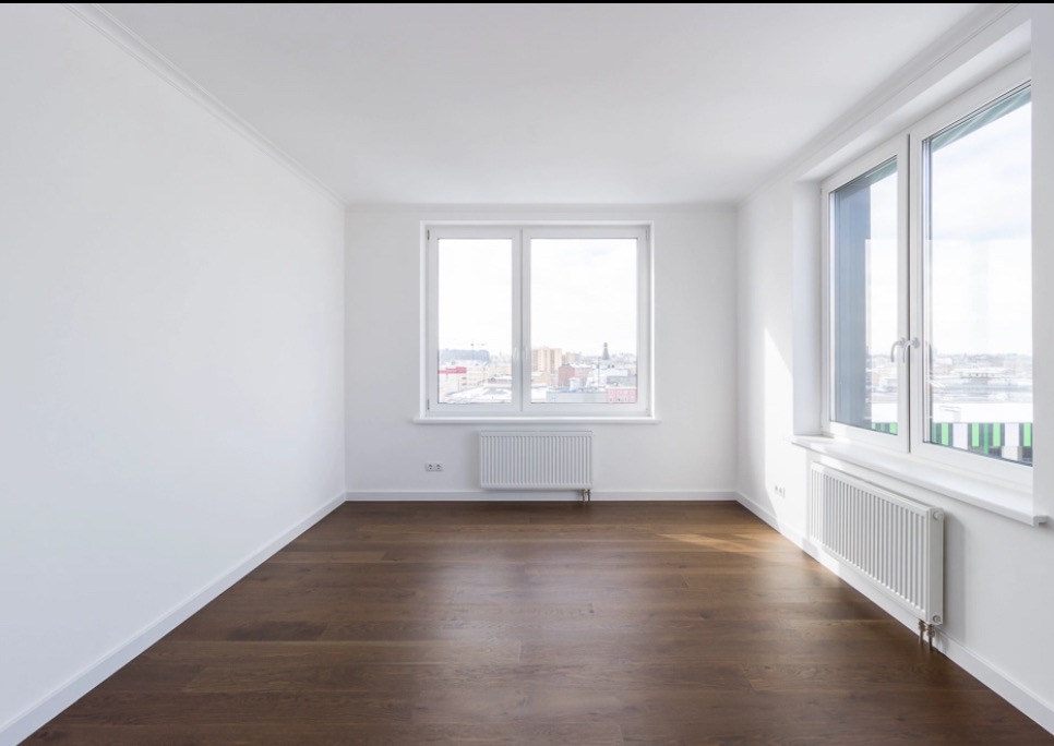 Jasne, puste wnętrze z drewnianą podłogą i białymi ścianami. Dwa okna z widokiem na miasto w tle. Minimalistyczny design, czyste linie i dużo naturalnego światła.