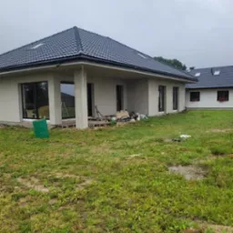 Dom jednorodzinny z ciemną dachówką karpiówką i szarymi ścianami zewnętrznymi. Widoczne narzędzia i materiały budowlane sugerują trwające prace wykończeniowe.