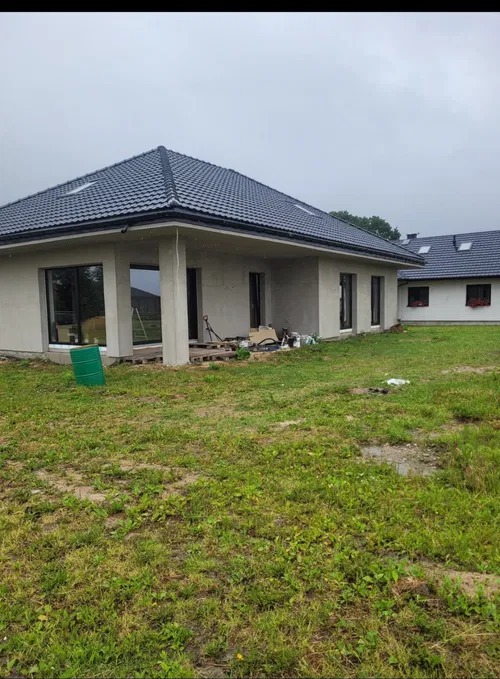 Dom jednorodzinny z ciemną dachówką karpiówką i szarymi ścianami zewnętrznymi. Widoczne narzędzia i materiały budowlane sugerują trwające prace wykończeniowe.