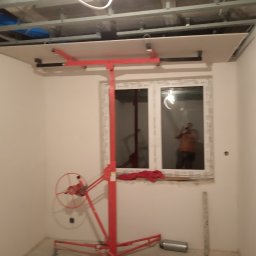 Tomasz Żabicki - Montaż płyty G-K na suficie za pomocą podnośnika. Widoczne metalowe profile konstrukcyjne i białe ściany. W oknie odbicie osoby robiącej zdjęcie.