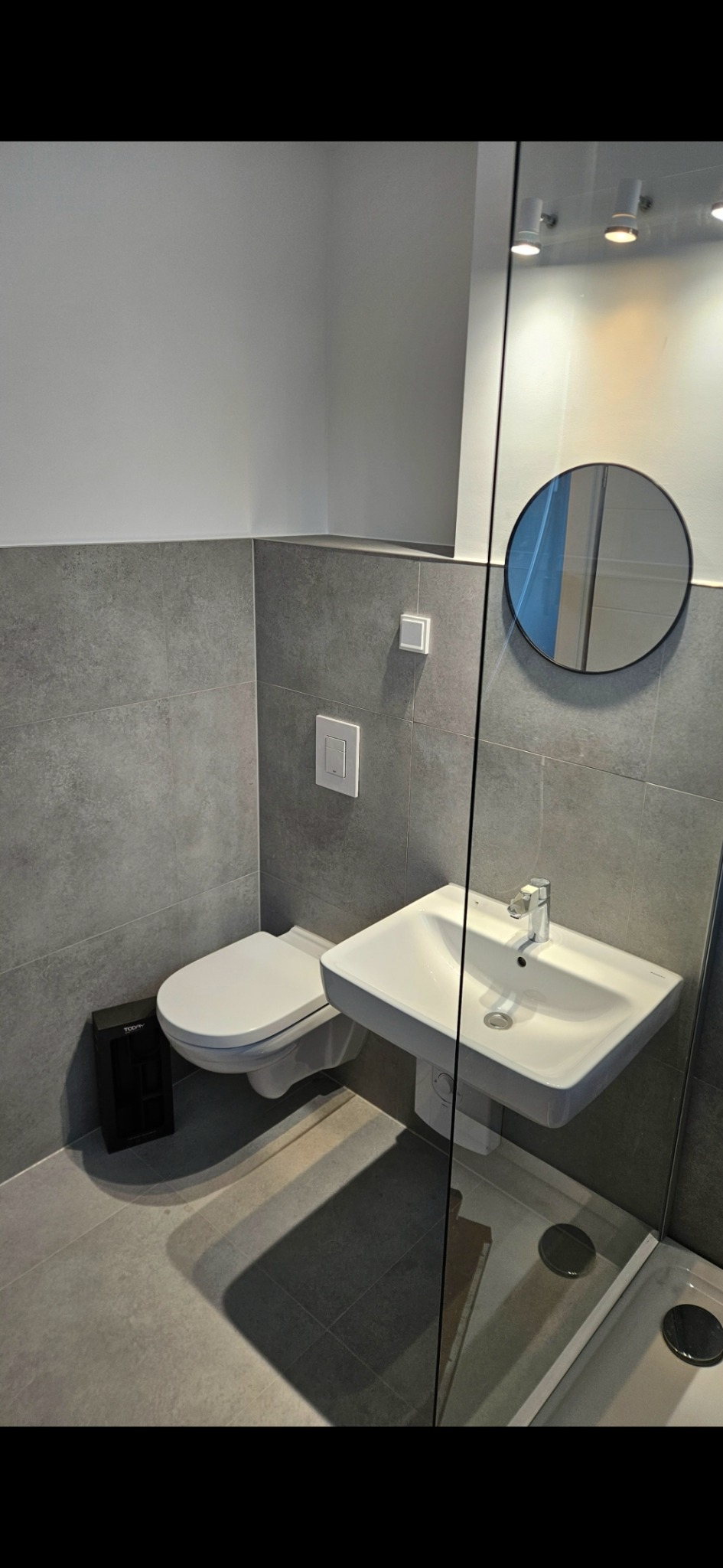 Nowoczesna łazienka w szarościach: wisząca toaleta, umywalka z baterią, okrągłe lustro i kabina prysznicowa ze szklaną ścianą. Minimalistyczny design.