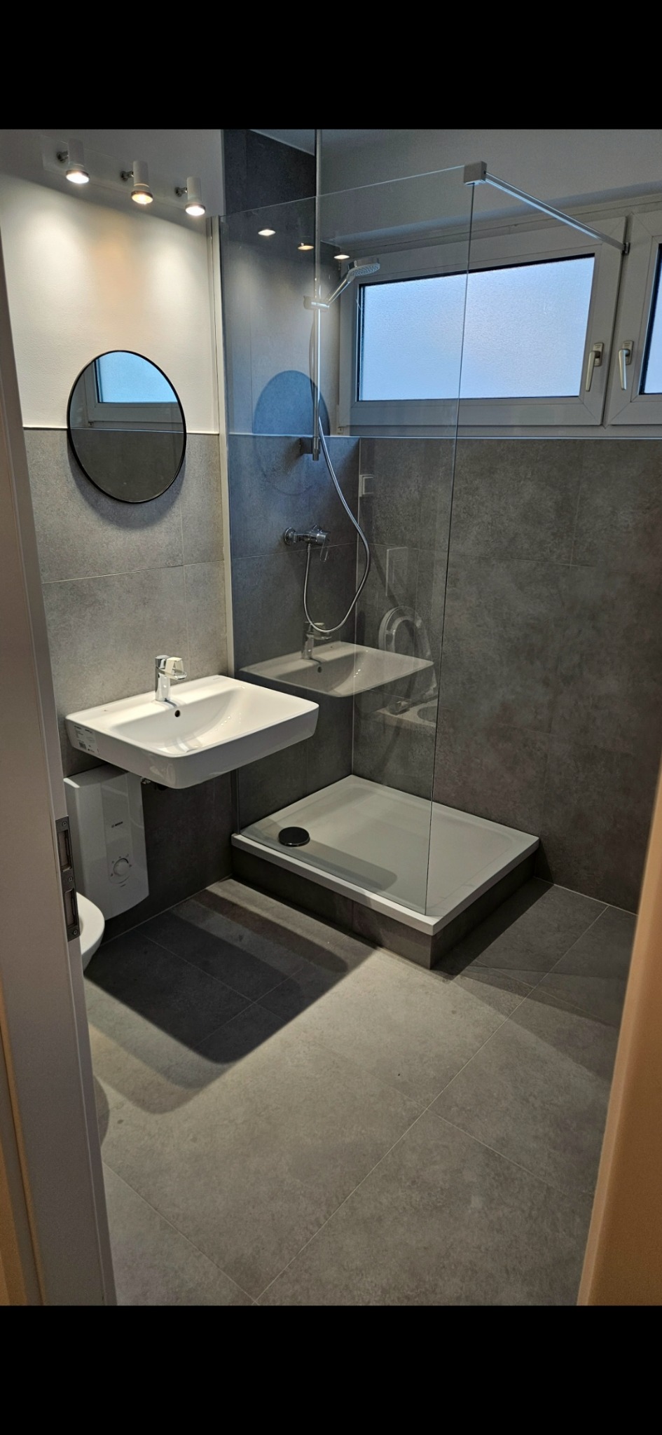 Nowoczesna łazienka w szarościach: kabina prysznicowa za szybą, okrągłe lustro nad umywalką, punktowe oświetlenie sufitowe i duże, grafitowe płytki podłogowe.