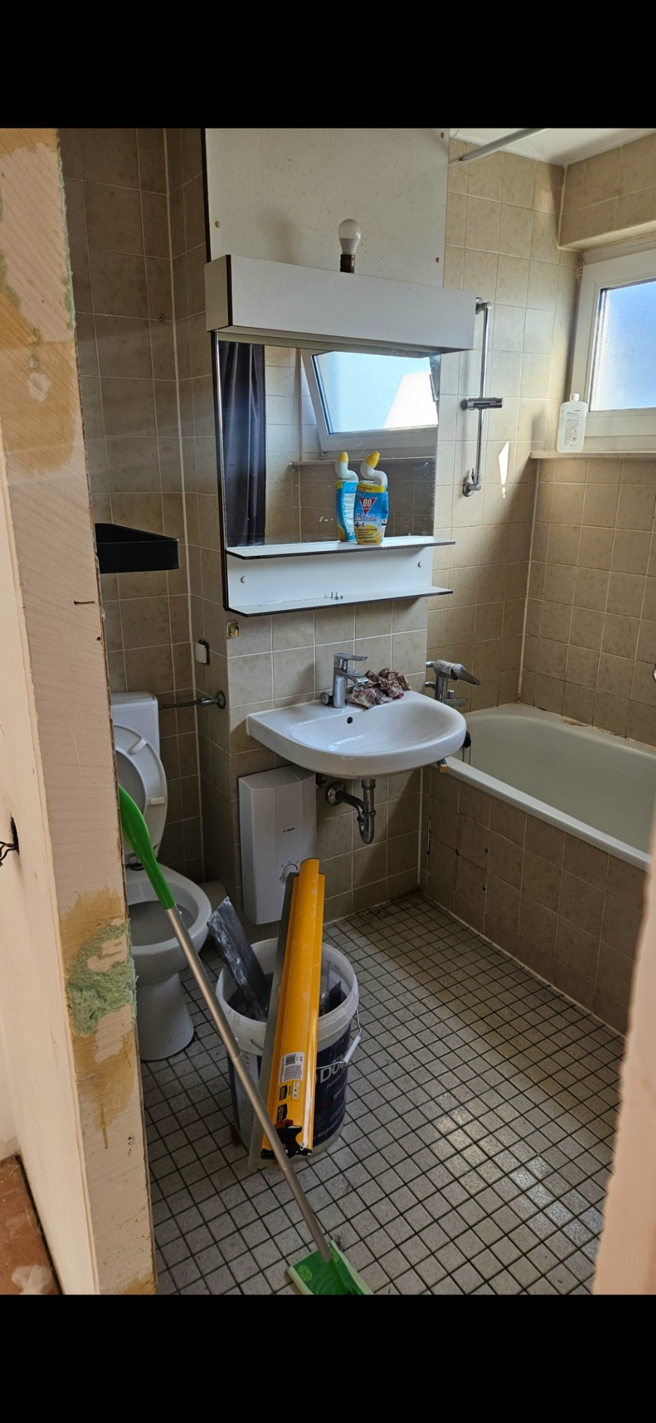 Łazienka w trakcie remontu: widok na umywalkę, wannę i narzędzia w wiadrze. Ściany wyłożone beżowymi płytkami, widoczne ślady po skuciu. Mop oparty o ścianę.