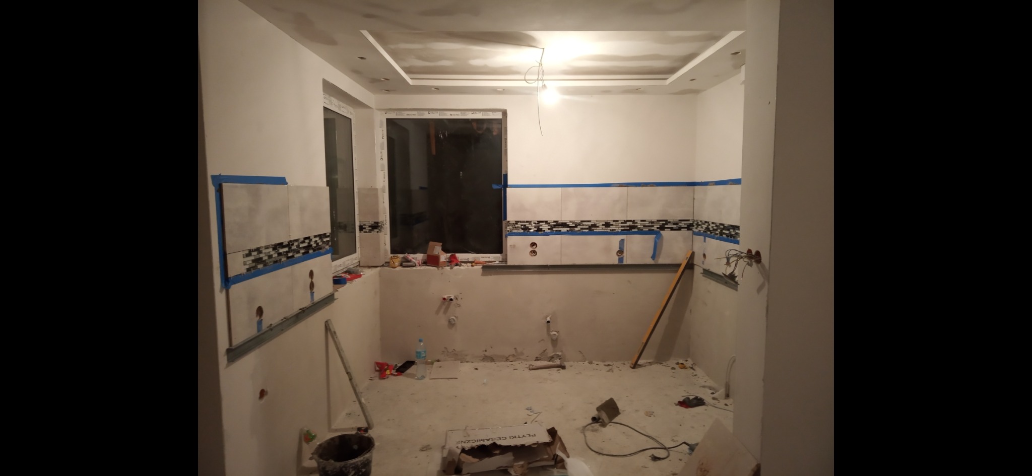 Wnętrze łazienki w trakcie remontu: częściowo położone płytki z dekoracyjnym pasem, widoczne instalacje, przygotowane ściany i sufit pod dalsze prace wykończeniowe.