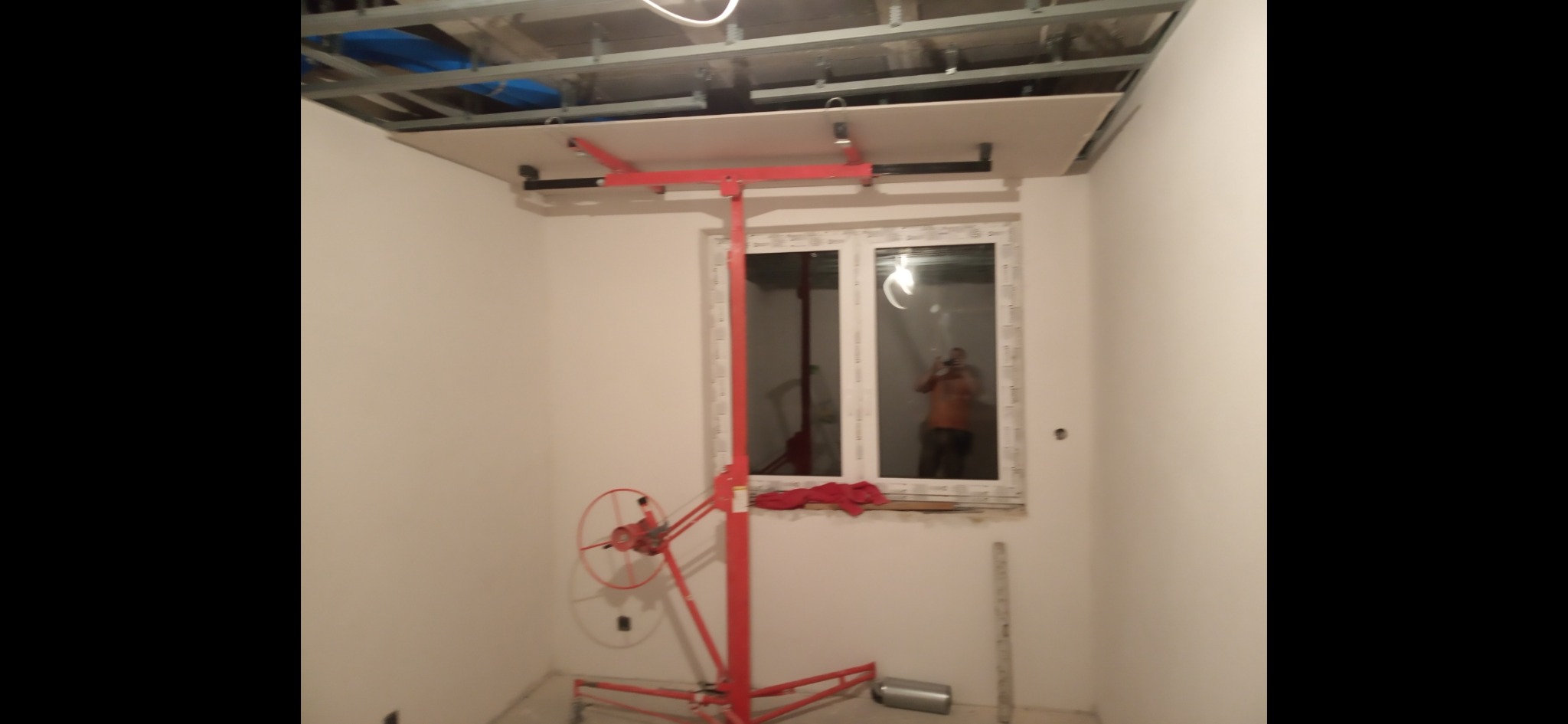 Montaż płyty G-K na suficie za pomocą podnośnika. Widoczne metalowe profile konstrukcyjne i białe ściany. W oknie odbicie osoby robiącej zdjęcie.