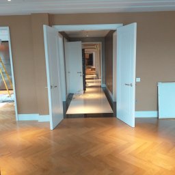 Firma - Nowoczesne wnętrze z parkietem w jodełkę i białymi drzwiami. Widok na długi korytarz z oświetleniem punktowym i białymi panelami. Ściany w kolorze beżowym.