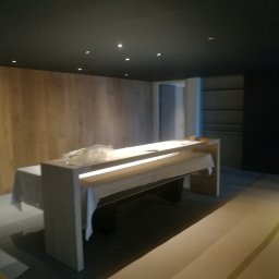 Firma - Nowoczesne wnętrze z drewnianą ścianą i minimalistycznym stołem z podświetleniem LED. Widoczne punkty świetlne na suficie i fragment osoby po lewej.