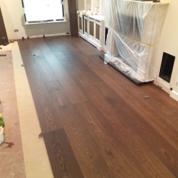 Firma - Układanie ciemnego parkietu w pokoju z oknem i osłoniętym kominkiem. Widoczny fragment podkładu podłogowego i narzędzia pracy. Parkiet o wyraźnym rysunku drewna.