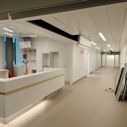 Firma - Nowoczesne, jasne wnętrze w trakcie wykańczania: białe ściany, recepcja z podświetleniem, widoczne kable i elementy instalacji. W tle okna i zabudowa meblowa.