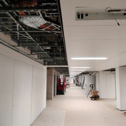 Firma - Długi korytarz w trakcie wykańczania z odsłoniętą instalacją pod sufitem. Widoczne kable, metalowe konstrukcje i częściowo zamontowany sufit podwieszany.