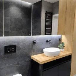 Nowoczesna łazienka z drewnianym blatem, umywalką nablatową i szafką w kolorze czarnym. Na ścianie szare płytki imitujące kamień i minimalistyczna armatura.