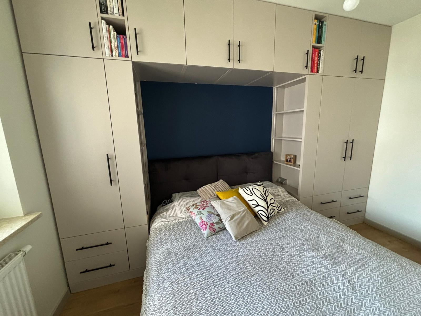 Jasna szafa wnękowa z półkami na książki, wkomponowana nad łóżkiem z wezgłowiem na tle granatowej ściany. Minimalistyczne, czarne uchwyty.