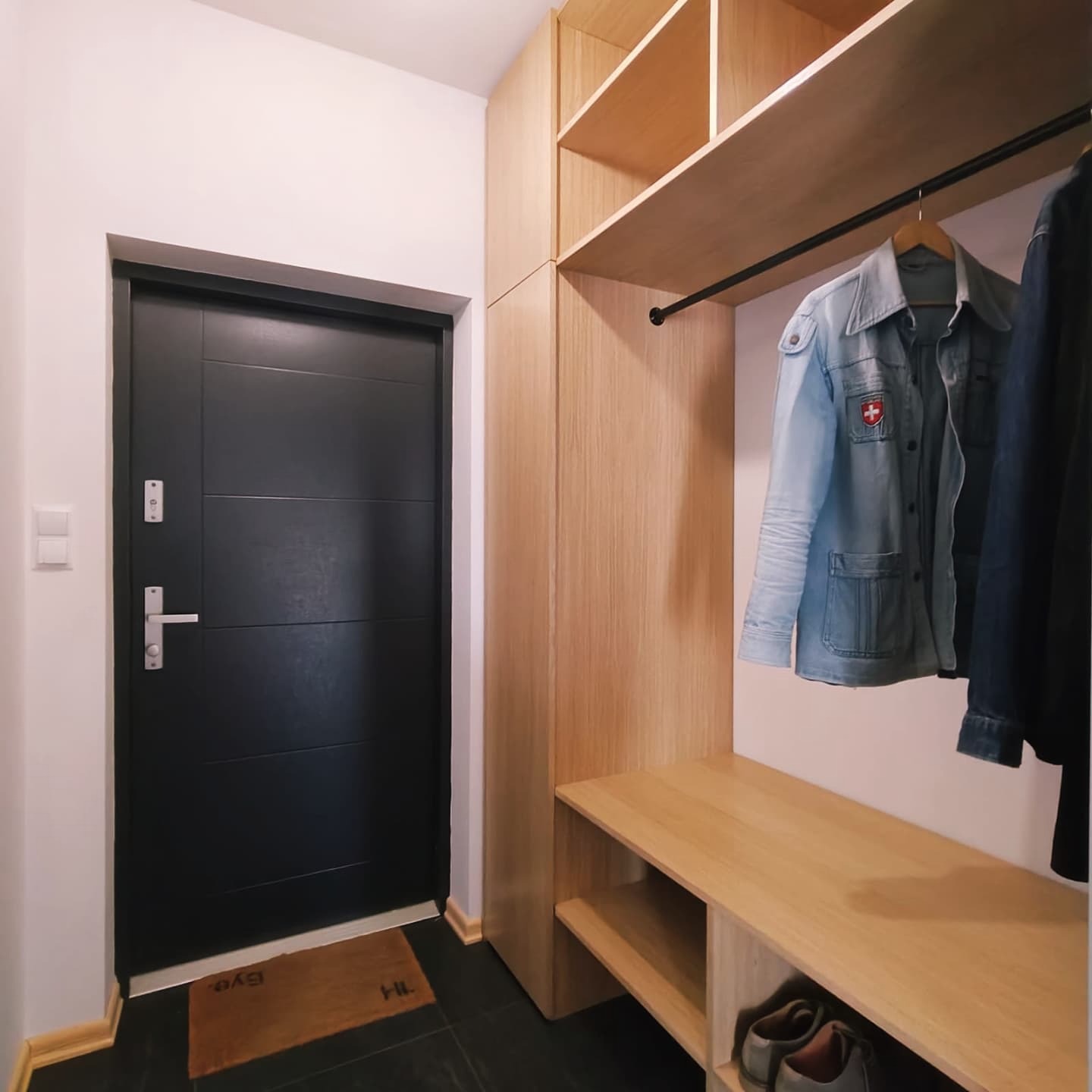 Nowoczesna szafa wnękowa z jasnego drewna w przedpokoju z czarną podłogą i drzwiami. Na wieszaku kurtka jeansowa.