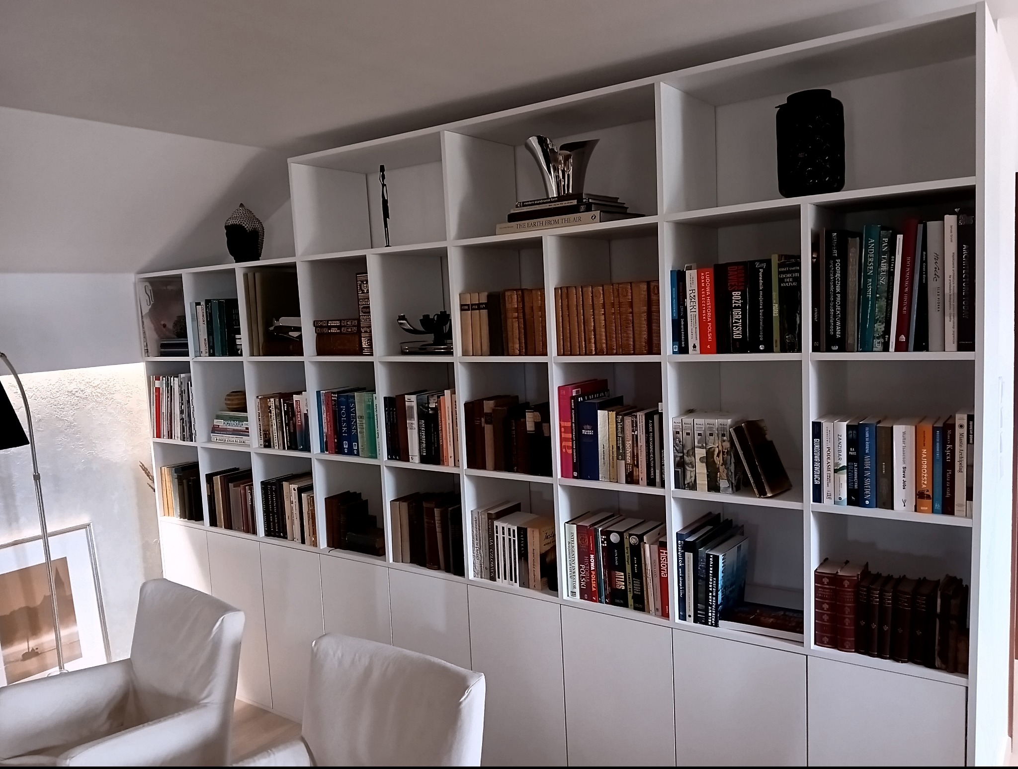 Biała, zabudowana regałowa szafa wnękowa z półkami wypełnionymi książkami i dekoracjami. W tle jasne ściany i dwa białe fotele. Subtelne oświetlenie.