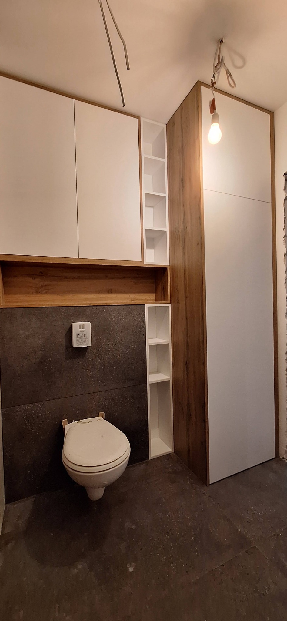 Nowoczesna zabudowa łazienkowa z szafkami w odcieniach bieli i drewna, z otwartymi półkami, nad wiszącą toaletą na tle grafitowych płytek. Surowy stan wykończenia podłogi.