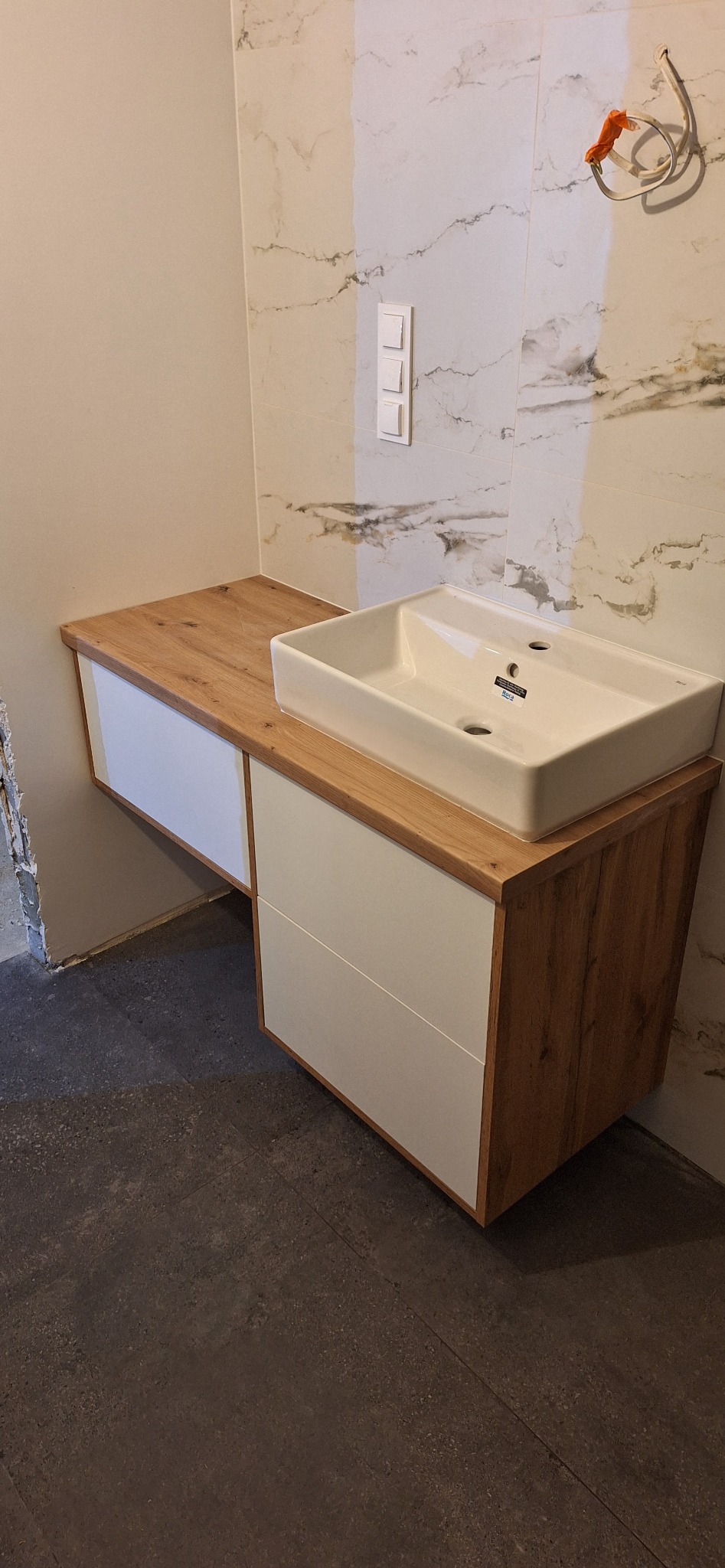 Nowoczesna szafka łazienkowa z blatem drewnopodobnym i białą umywalką prostokątną. Widoczne płytki imitujące marmur. Surowy stan wykończenia pomieszczenia.