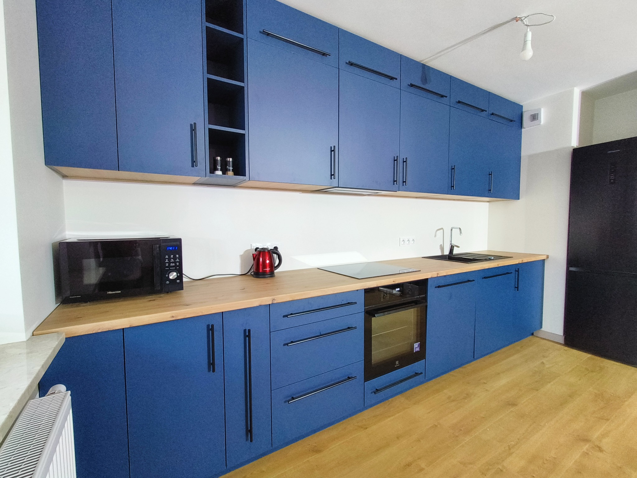 Niebieska zabudowa kuchenna z drewnianym blatem, czarnymi uchwytami i sprzętami AGD. Górne szafki sięgają sufitu, a na blacie stoi czajnik i mikrofalówka.