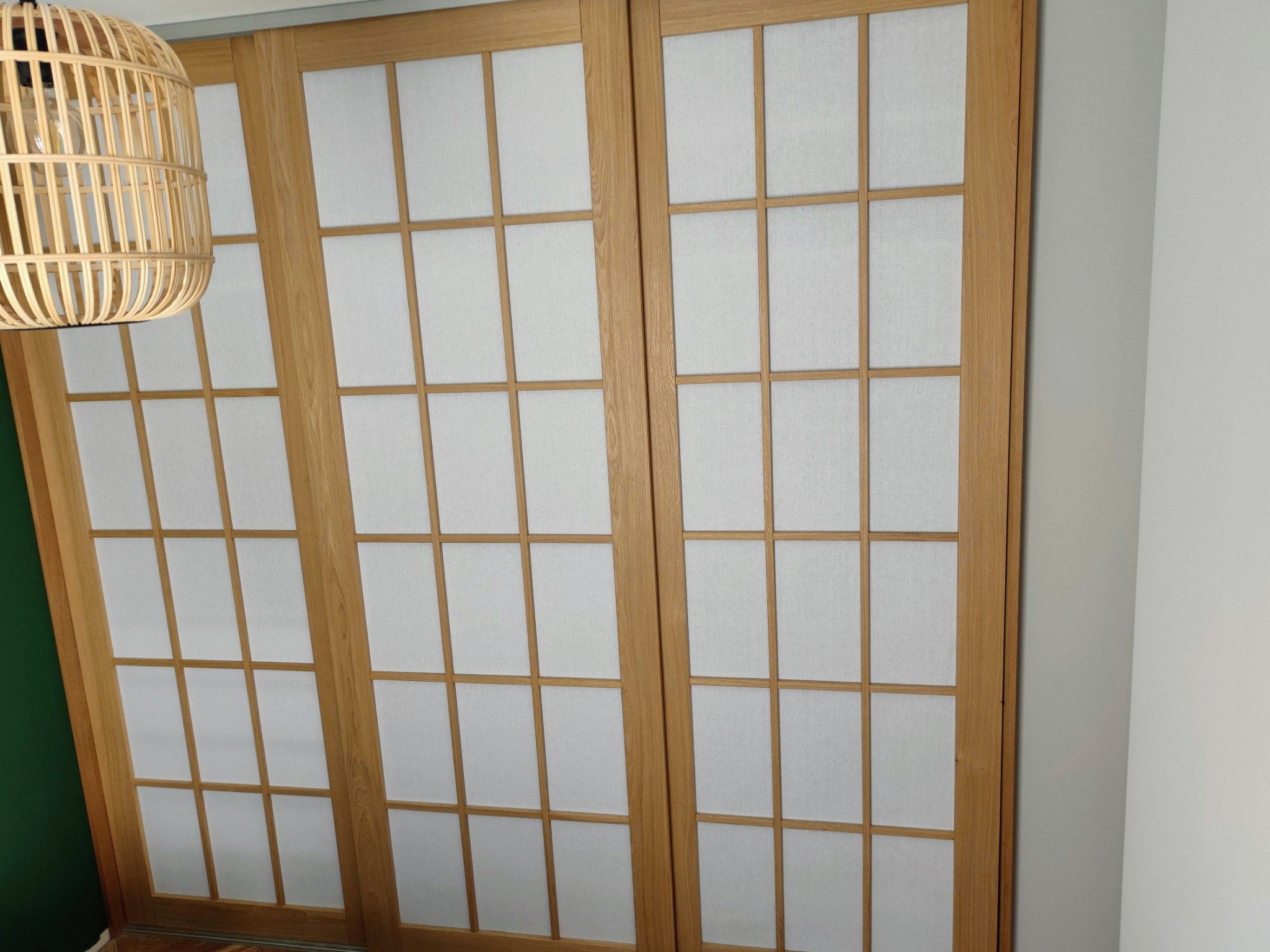 Drewniane, przesuwne drzwi z białymi panelami w stylu japońskim, oświetlone wiszącą lampą z naturalnego rattanu. Widoczny fragment zielonej ściany.