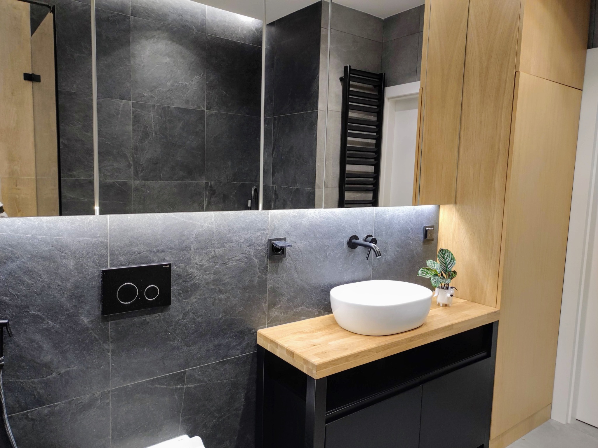 Nowoczesna łazienka z drewnianym blatem, umywalką nablatową i szafką w kolorze czarnym. Na ścianie szare płytki imitujące kamień i minimalistyczna armatura.