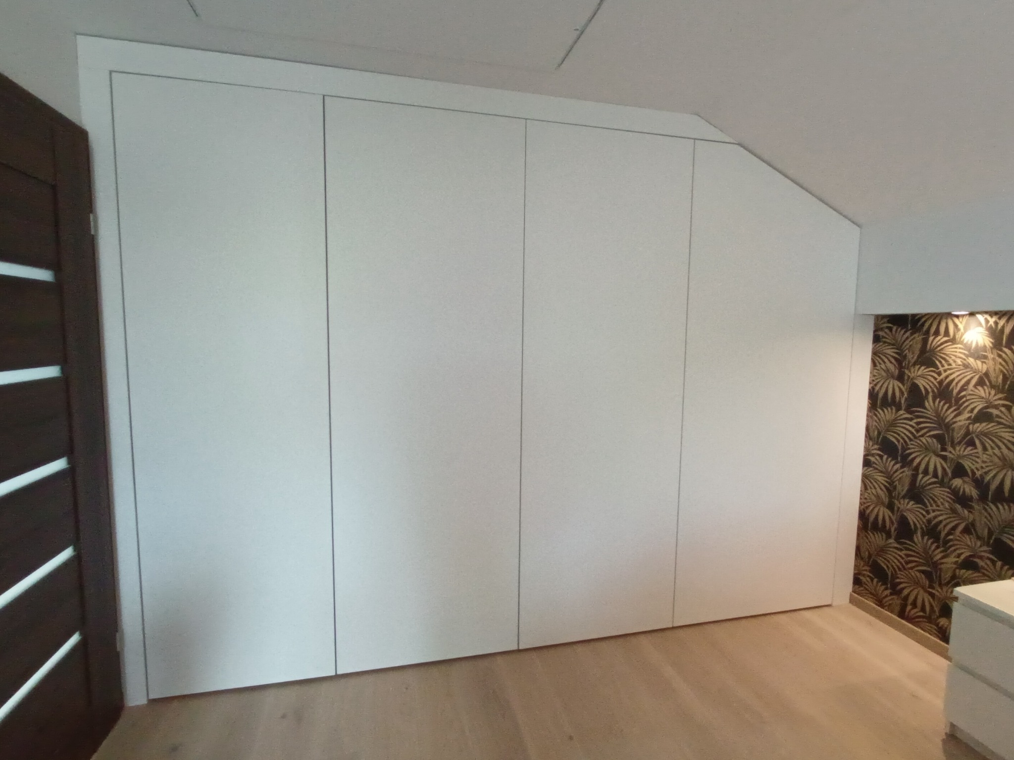 Biała szafa wnękowa z czterema drzwiami, dopasowana do skosu dachu, z widocznym fragmentem drewnianej podłogi i drzwi.