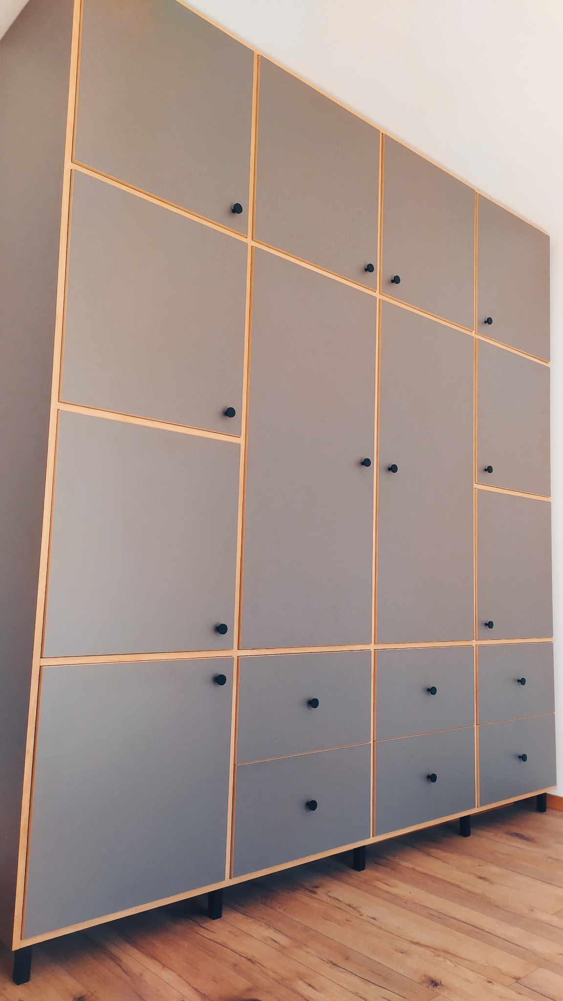 Szara szafa na wymiar z drewnianymi obramowaniami i czarnymi gałkami, ustawiona na niskich nóżkach, na tle drewnianej podłogi i białej ściany.