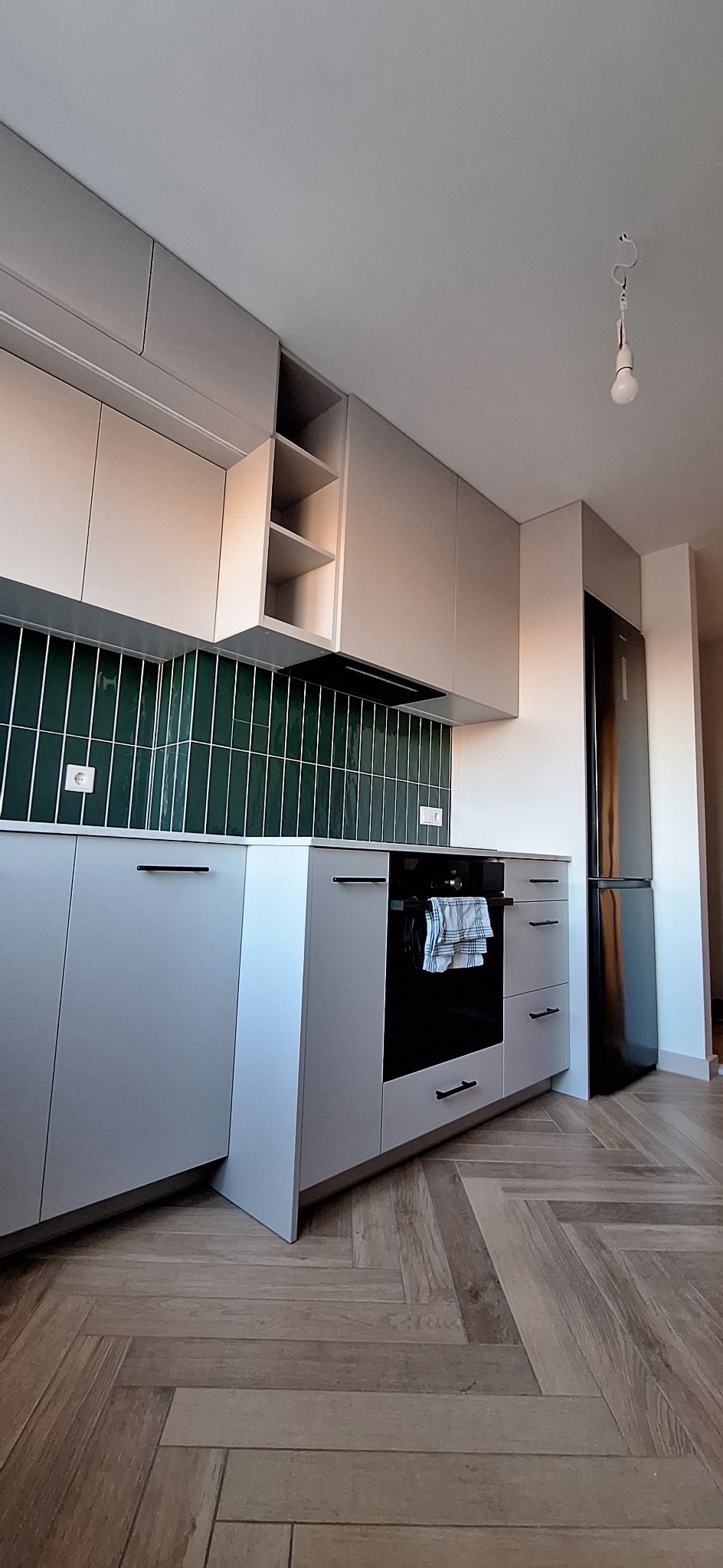 Szara kuchnia na wymiar z zielonymi płytkami i drewnianą podłogą w jodełkę. Widoczny piekarnik z ręcznikiem i czarna lodówka. Oświetlenie sufitowe.