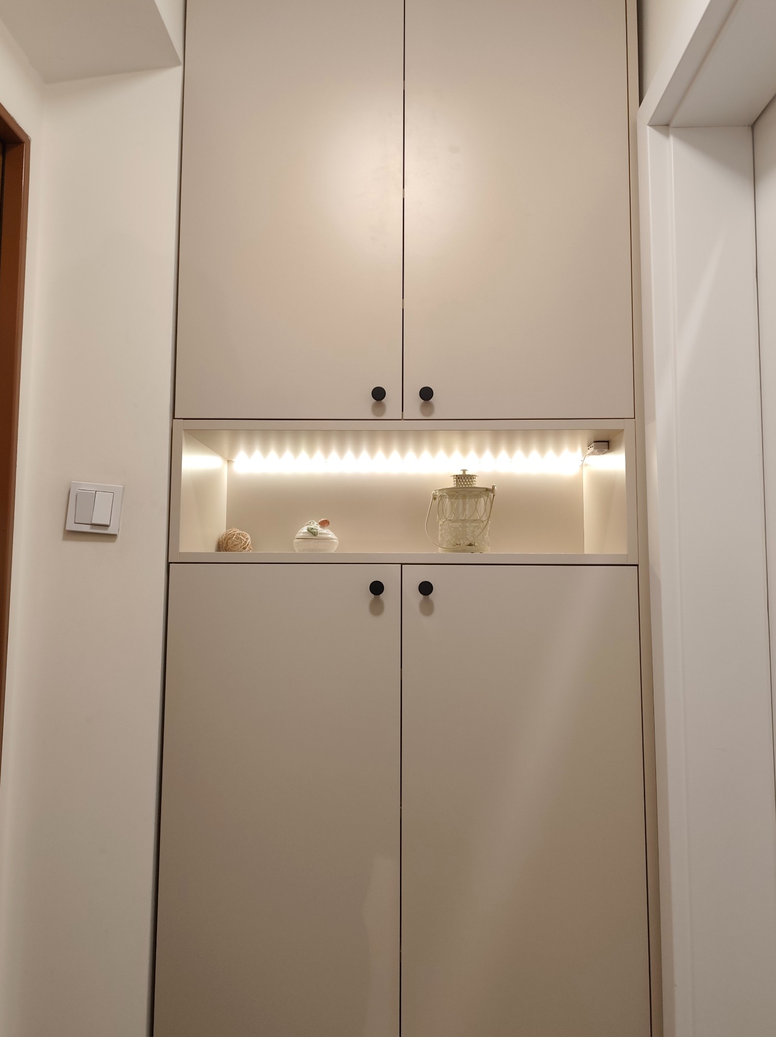 Beżowa szafa wnękowa z oświetleniem LED półki, czarne okrągłe uchwyty. Dekoracje na półce: szklany lampion, ceramiczna figurka i kula z włóczki. Minimalistyczny design.