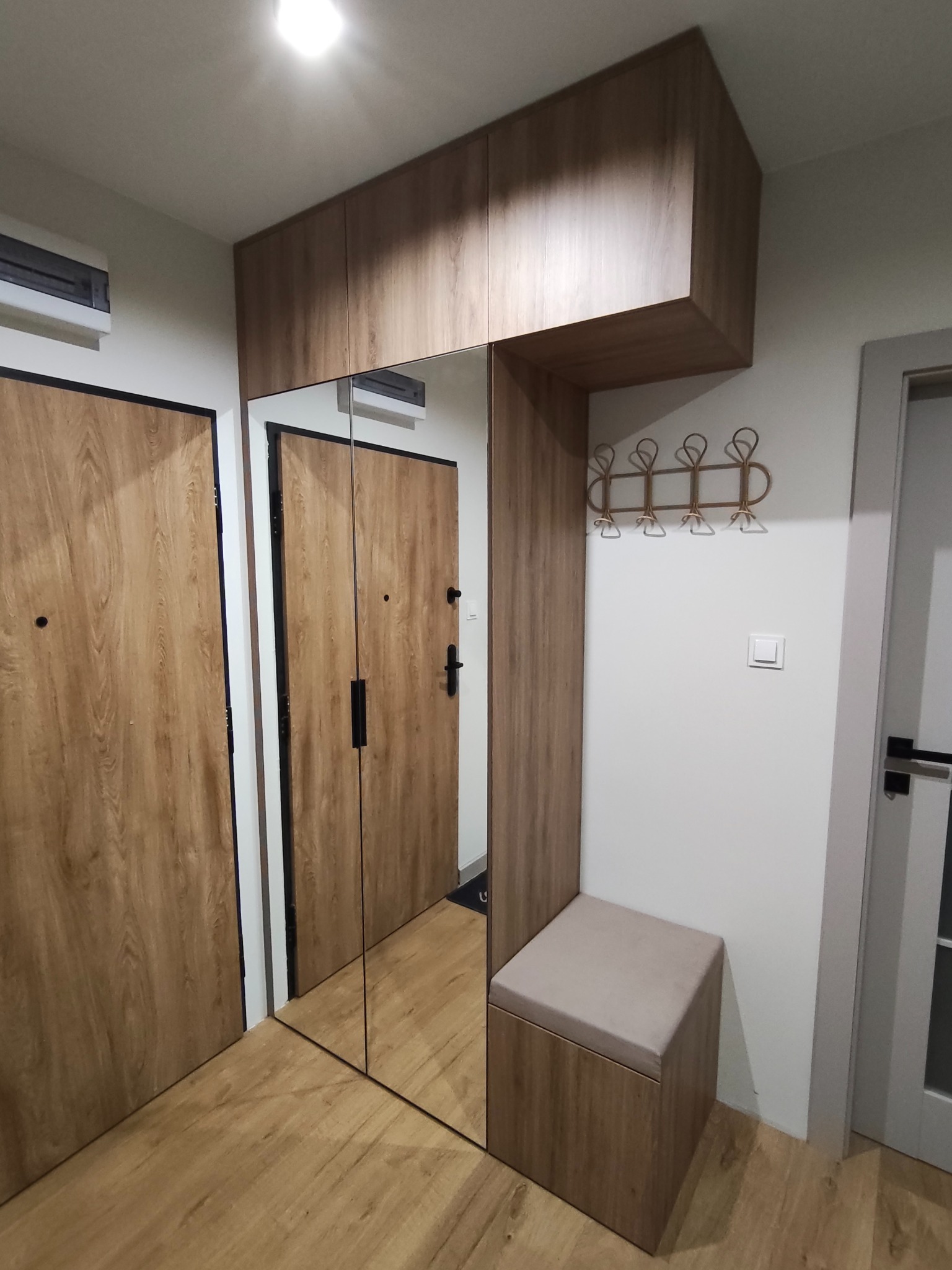 Funkcjonalna zabudowa przedpokoju z lustrem, szafkami i siedziskiem w odcieniach drewna. W tle widoczne drzwi wejściowe i podłoga w podobnej kolorystyce.