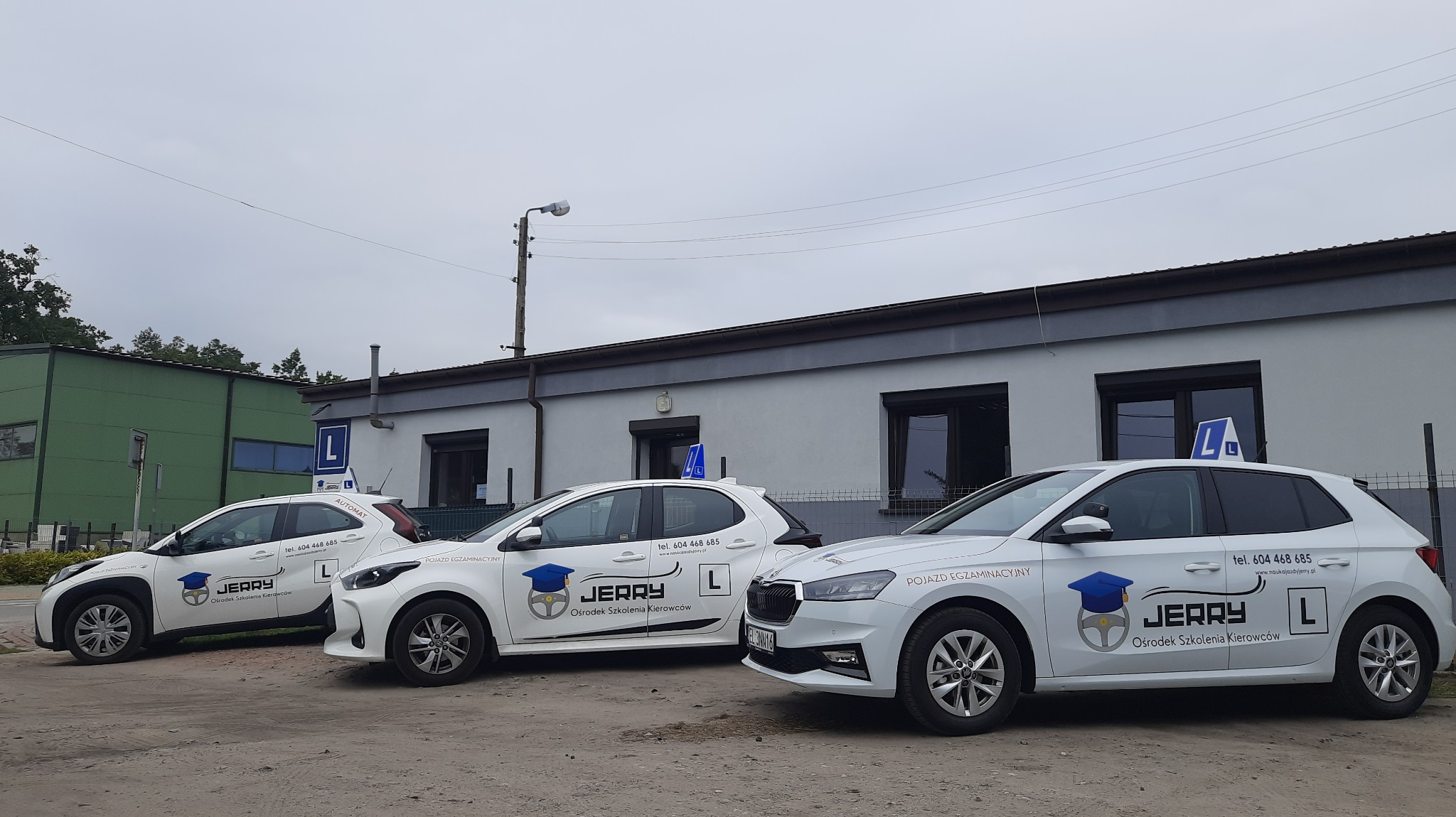 Trzy białe samochody z logo szkoły jazdy 'Jerry' zaparkowane przed budynkiem. Na dachu każdego auta widnieje niebieska litera 'L'. Pochmurne niebo w tle.