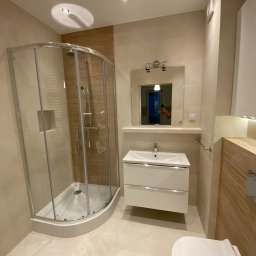 LevelProBud - Jasna łazienka po remoncie: kabina prysznicowa z zaokrąglonym szkłem, biała umywalka z szafką, toaleta. Beżowe płytki imitujące drewno na ścianach i podłodze. Nowoczesne oświetlenie.