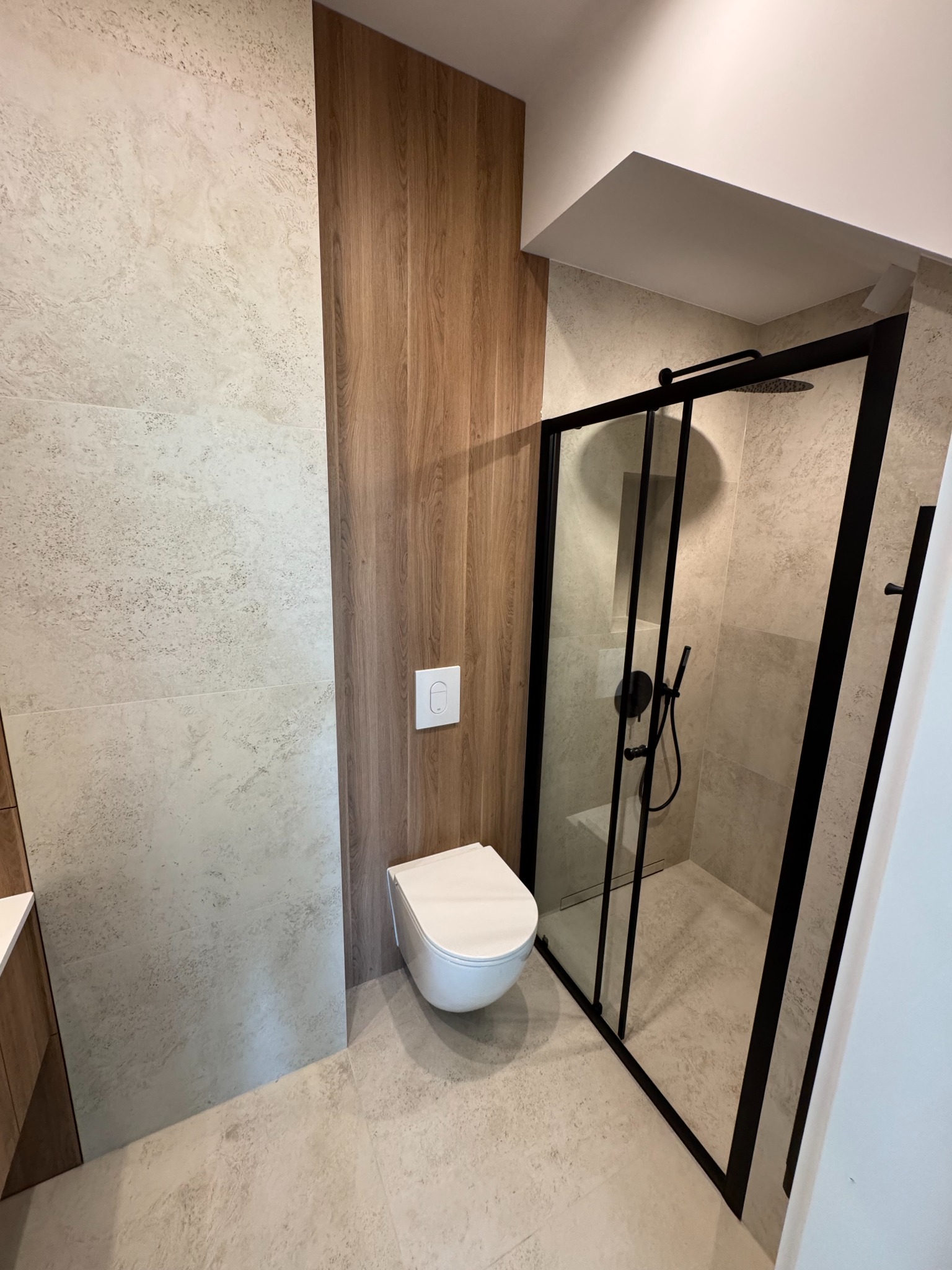 Nowoczesna łazienka po remoncie: kabina prysznicowa z czarną ramą, podwieszana toaleta, beżowe płytki imitujące kamień i drewnopodobna okładzina ściany.