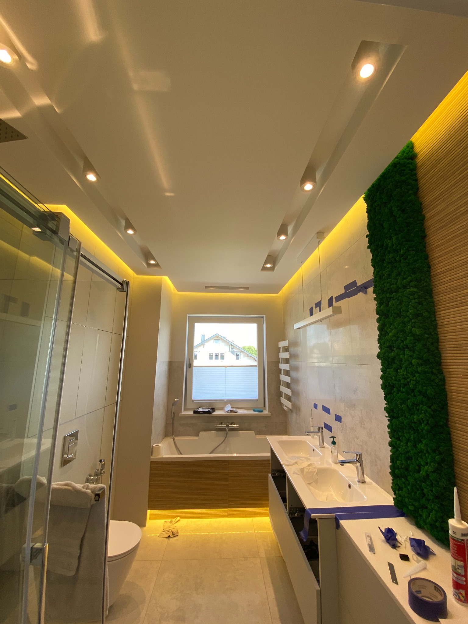 Nowoczesna łazienka z podświetleniem LED, wanną, umywalką i pionową ścianą z mchu. Widoczne narzędzia sugerują prace wykończeniowe.