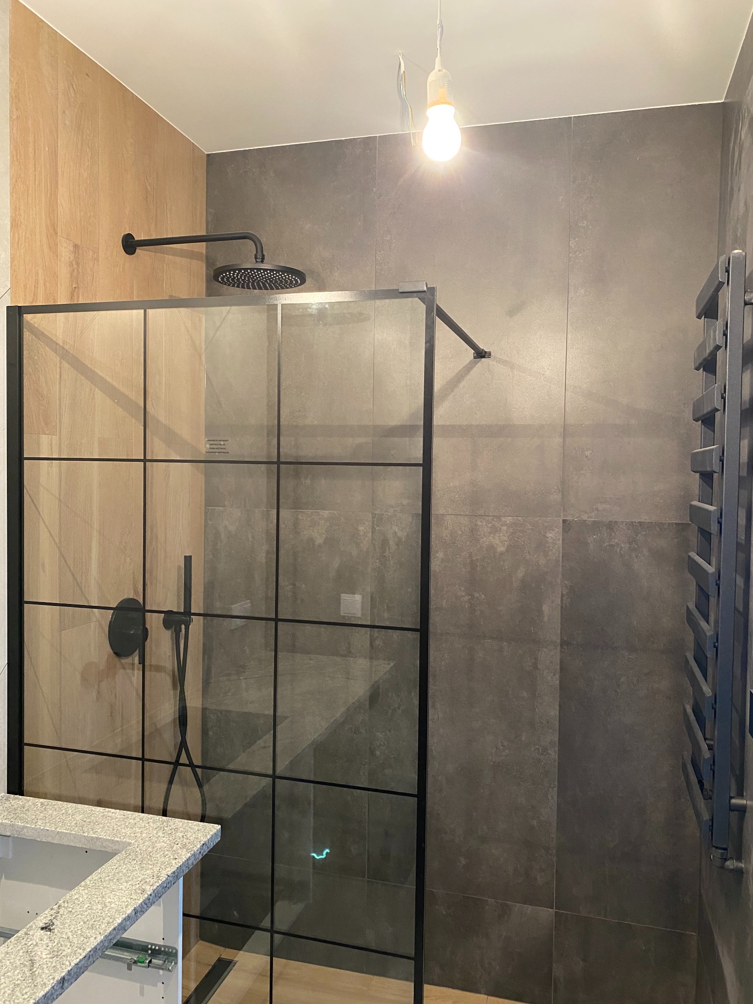 Nowoczesna kabina prysznicowa z czarnymi profilami i deszczownicą, szare płytki, drewnopodobne panele, grzejnik drabinkowy w łazience po remoncie.