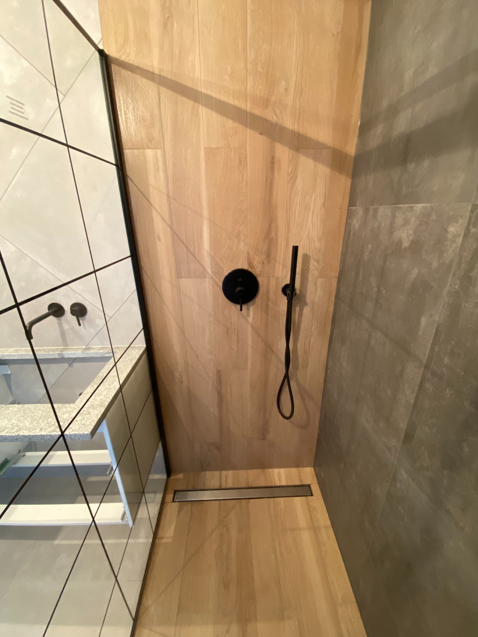 Nowoczesna kabina prysznicowa z drewnopodobną okładziną, czarną armaturą i odpływem liniowym. Widoczna umywalka z szafką w tle.