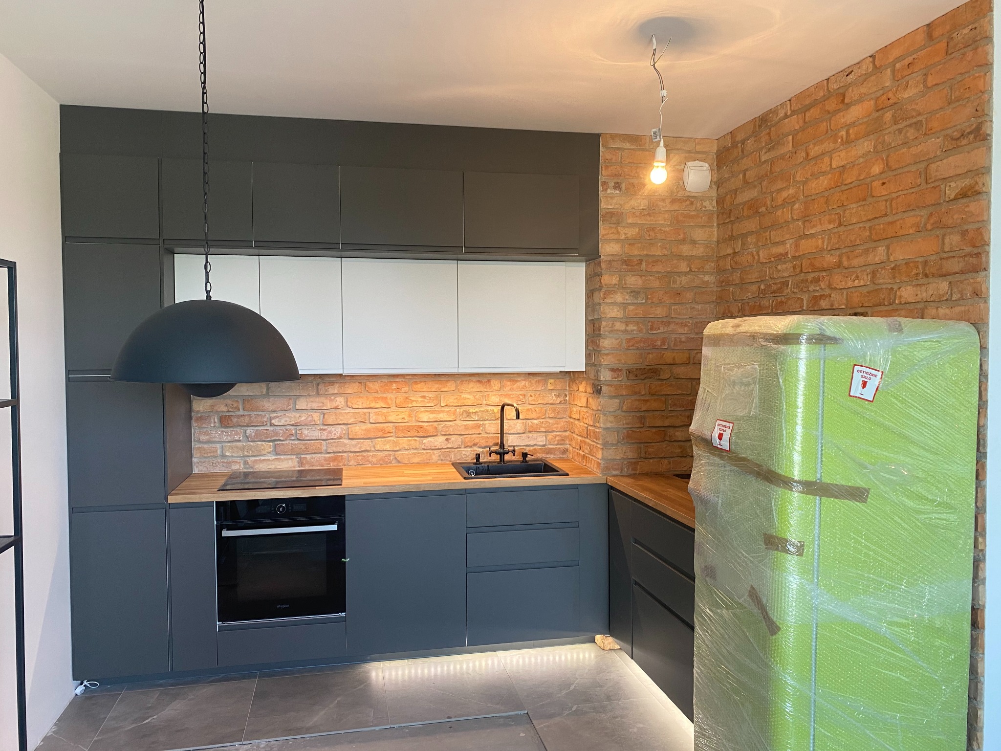 Nowoczesna kuchnia z szarymi szafkami, ceglaną ścianą i drewnianym blatem. W tle lodówka zabezpieczona folią. Minimalistyczny design i oświetlenie punktowe.