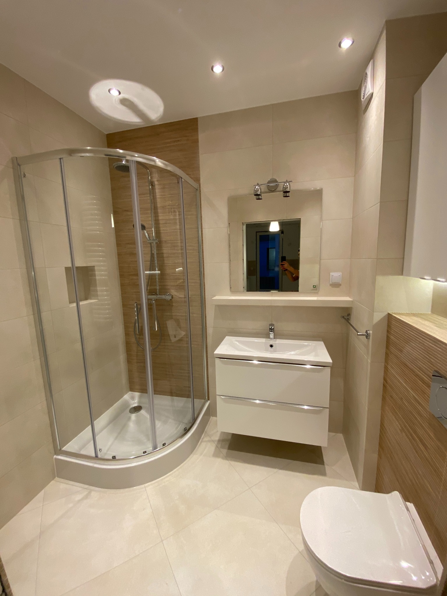 Jasna łazienka po remoncie: kabina prysznicowa z zaokrąglonym szkłem, biała umywalka z szafką, toaleta. Beżowe płytki imitujące drewno na ścianach i podłodze. Nowoczesne oświetlenie.