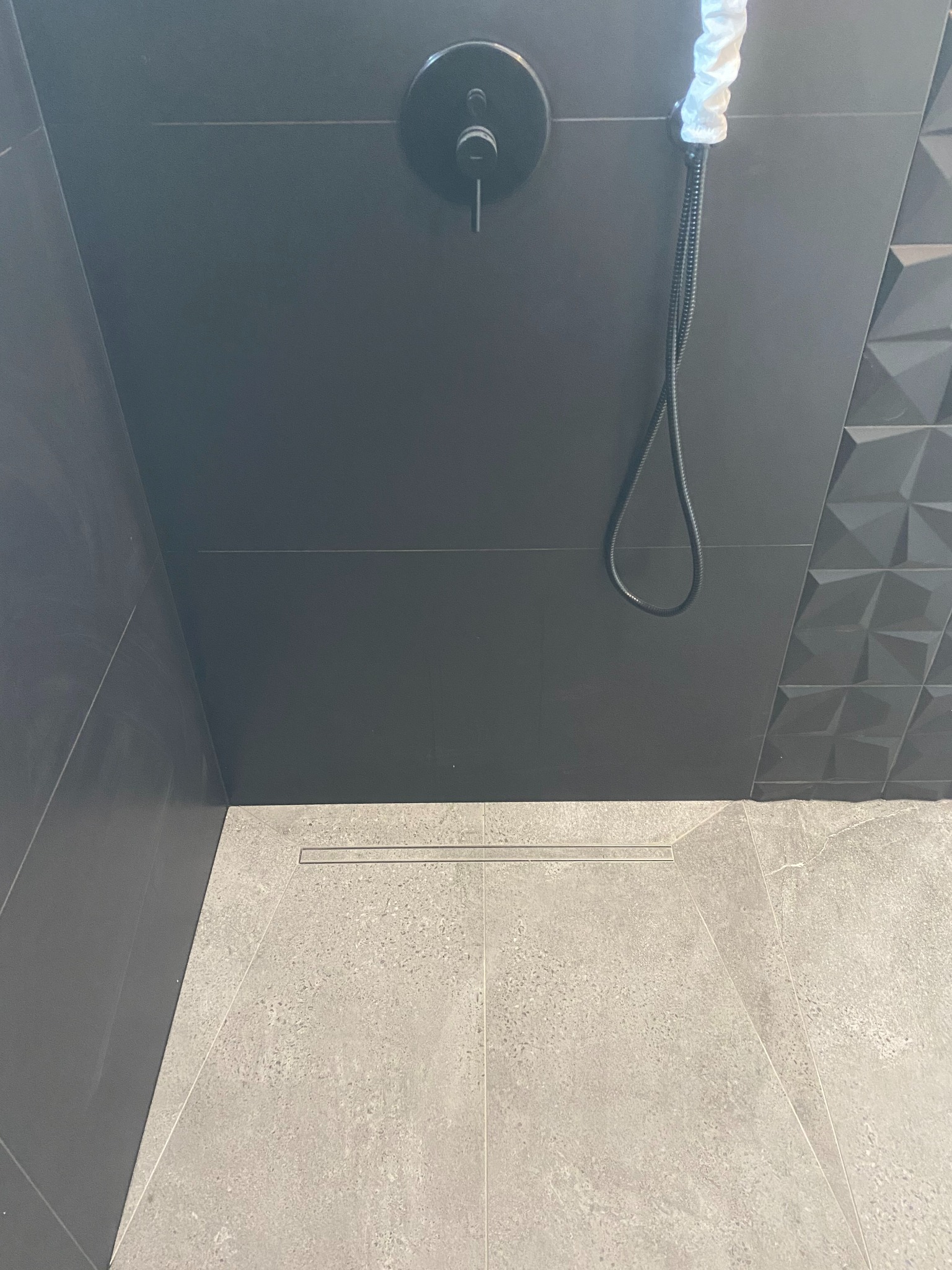 Nowoczesna kabina prysznicowa z czarną armaturą i szarymi płytkami, w tym trójwymiarowy panel dekoracyjny. Widoczny odpływ liniowy i ochrona głowicy prysznicowej.