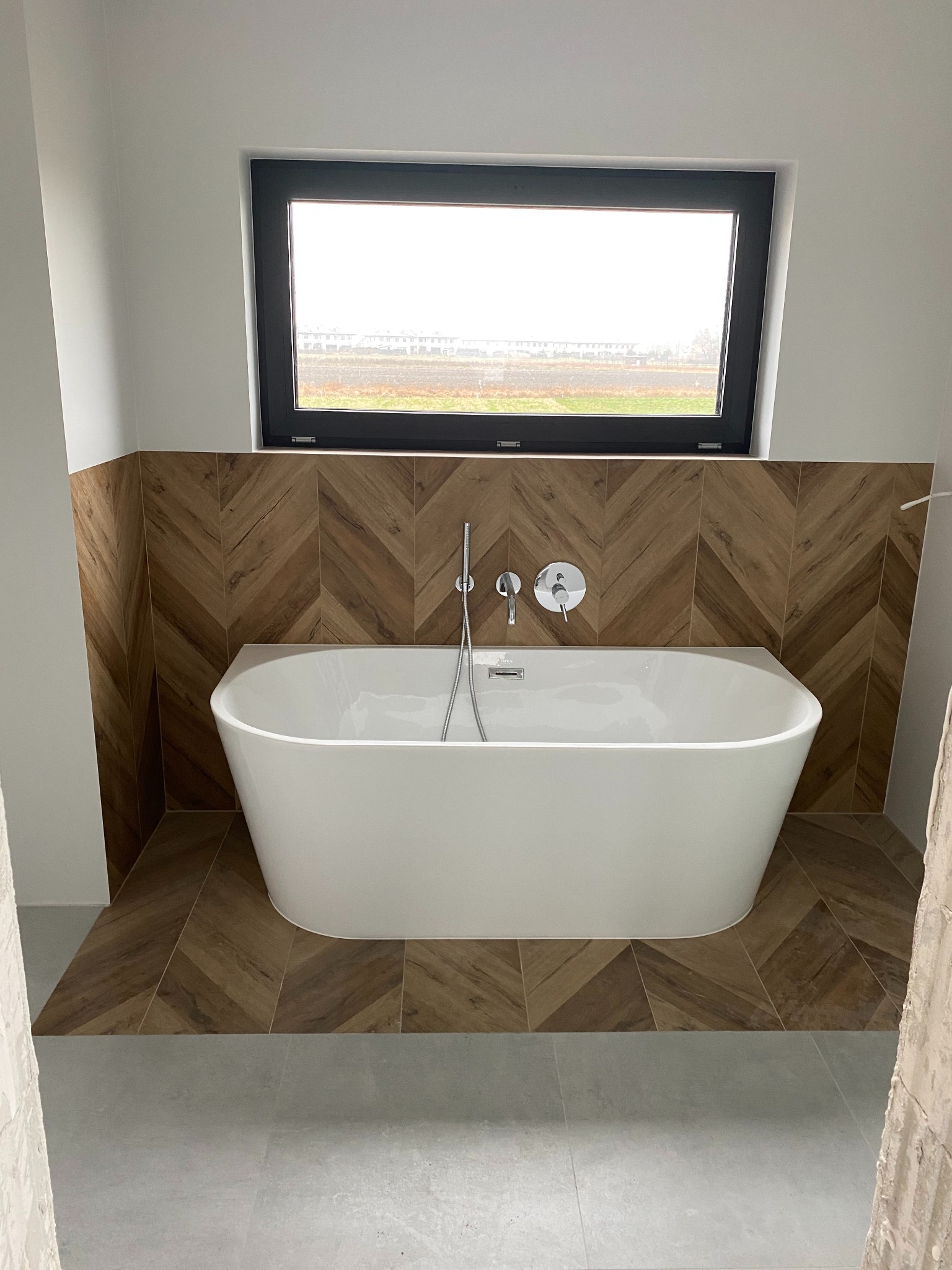 Nowoczesna łazienka z wolnostojącą wanną, drewnopodobnymi płytkami ułożonymi w jodełkę na ścianie i podłodze, oraz dużym oknem z widokiem na zewnątrz.