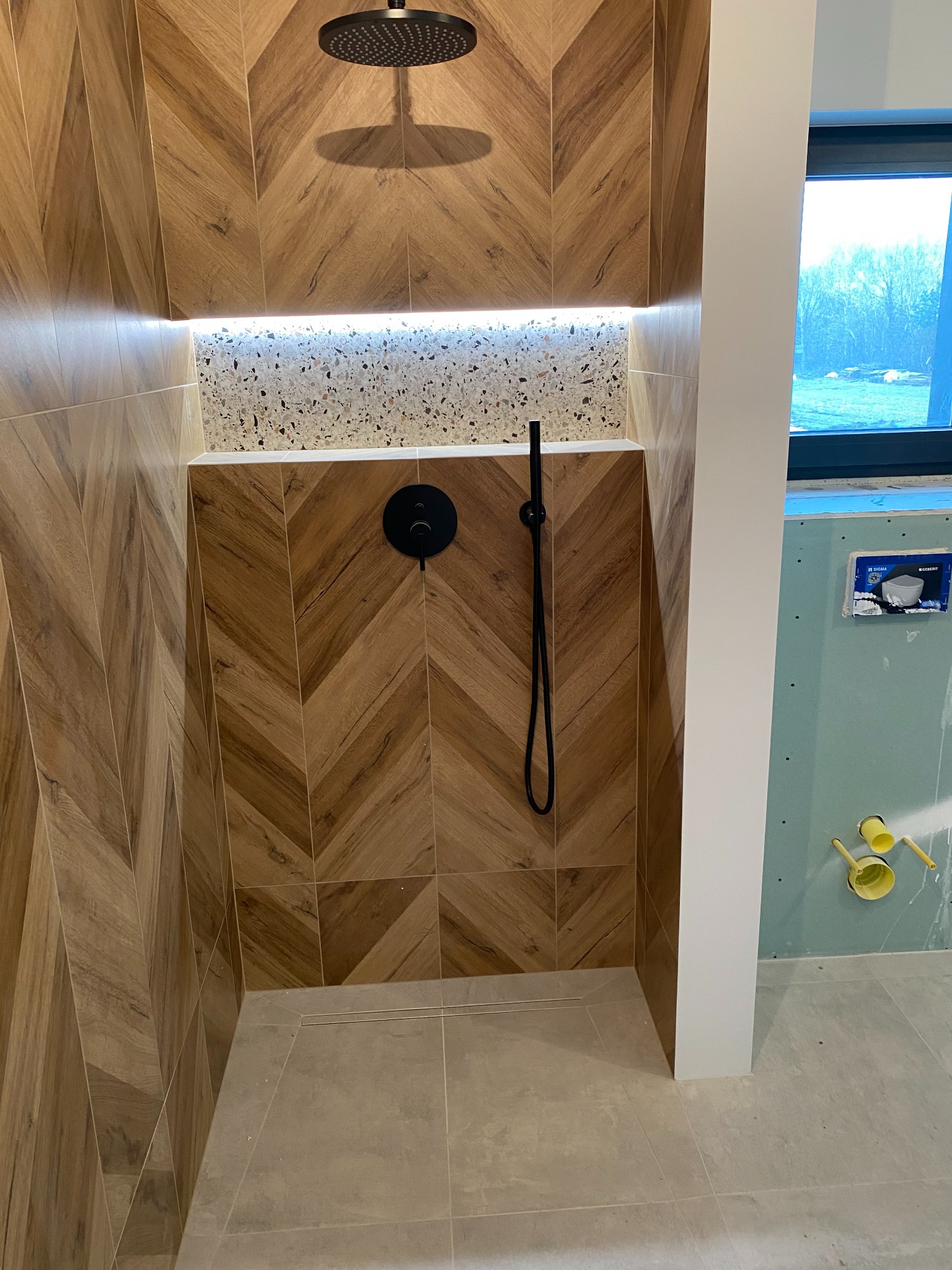 Nowoczesna kabina prysznicowa z drewnopodobnymi płytkami ułożonymi w jodełkę, czarną armaturą i wnęką z mozaiką, częściowo wykończona, z widoczną instalacją.