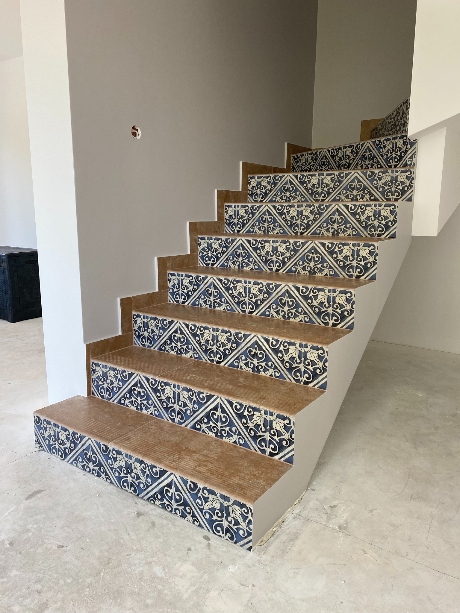 Schody wykończone dekoracyjnymi, wzorzystymi płytkami w odcieniach brązu i granatu. Widoczne detale wykończenia, surowy beton na podłodze i otwór w ścianie na instalację.