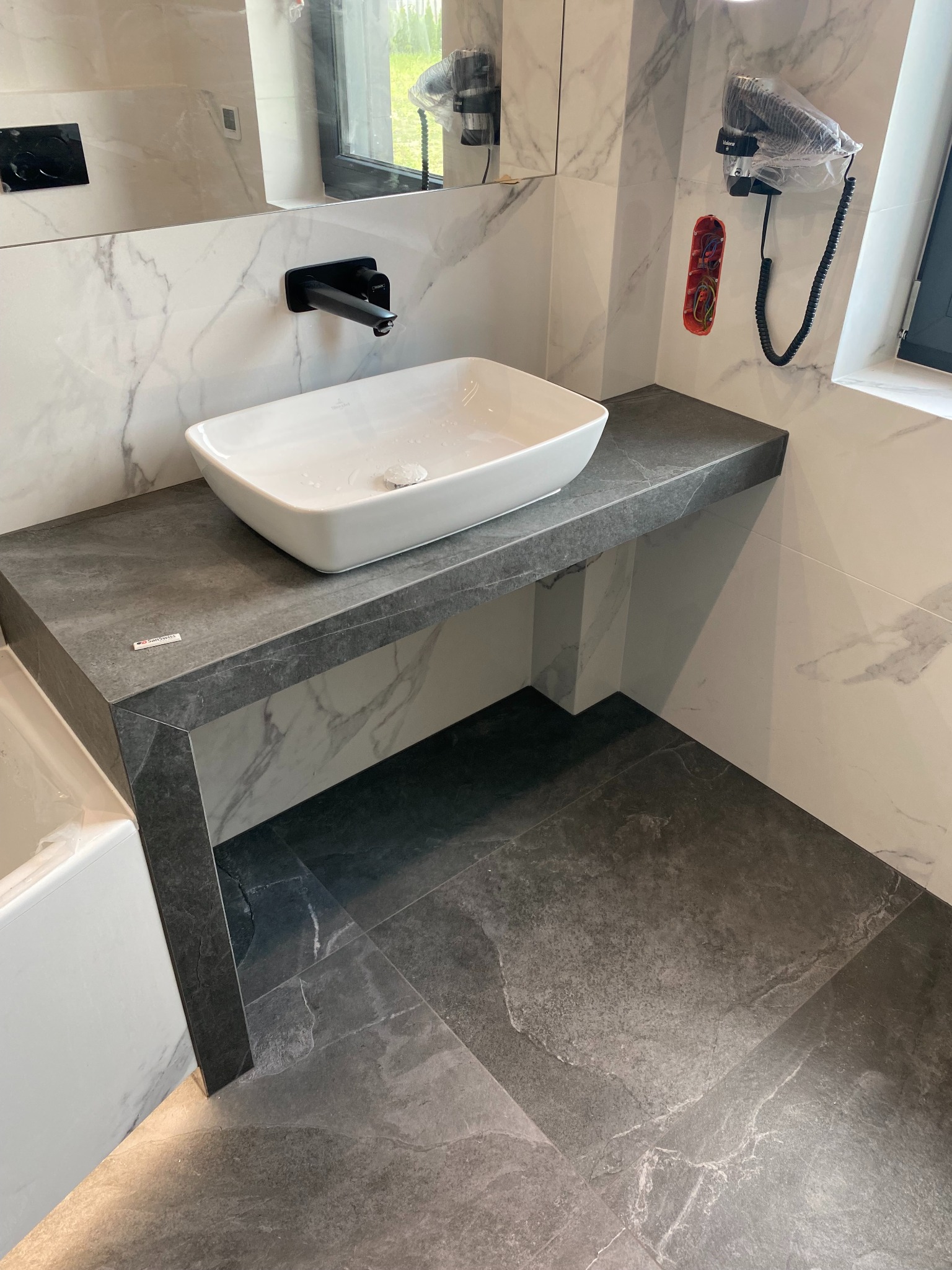 Biała, prostokątna umywalka na szarym blacie z czarną armaturą. Marmurowe ściany i podłoga z szarych płytek. Widoczny fragment wanny i suszarka na ścianie.