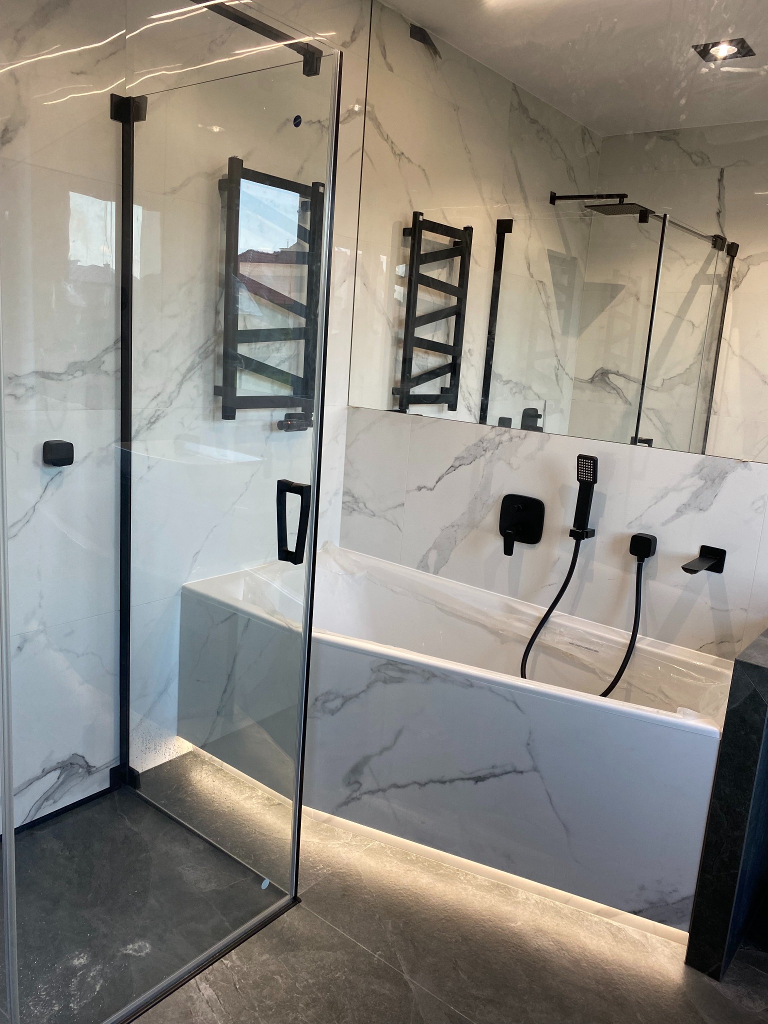 Elegancka łazienka z kabiną prysznicową typu walk-in, białą wanną i marmurowymi płytkami. Czarne akcenty armatury i grzejnika dodają nowoczesnego charakteru.