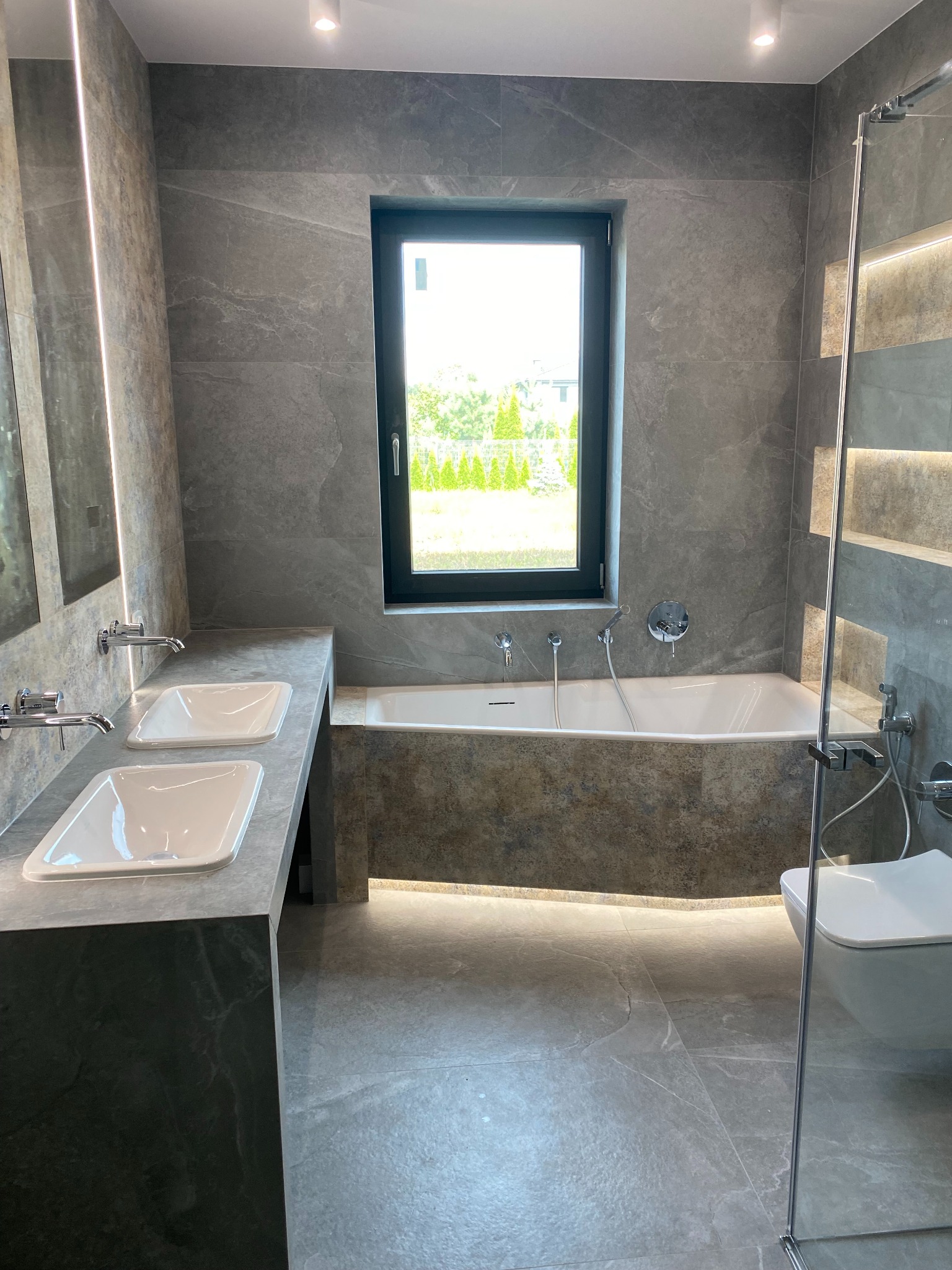 Nowoczesna łazienka w szarościach z podświetlaną wanną, dwiema umywalkami i dużym oknem z widokiem na zieleń, podkreślona minimalistycznym designem i stonowaną kolorystyką.
