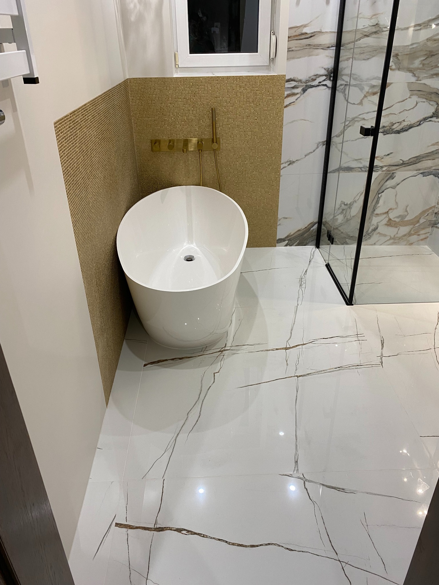 Nowoczesna łazienka z wolnostojącą wanną, złote detale armatury i mozaiki, białe płytki imitujące marmur, kabina prysznicowa z czarnymi profilami.