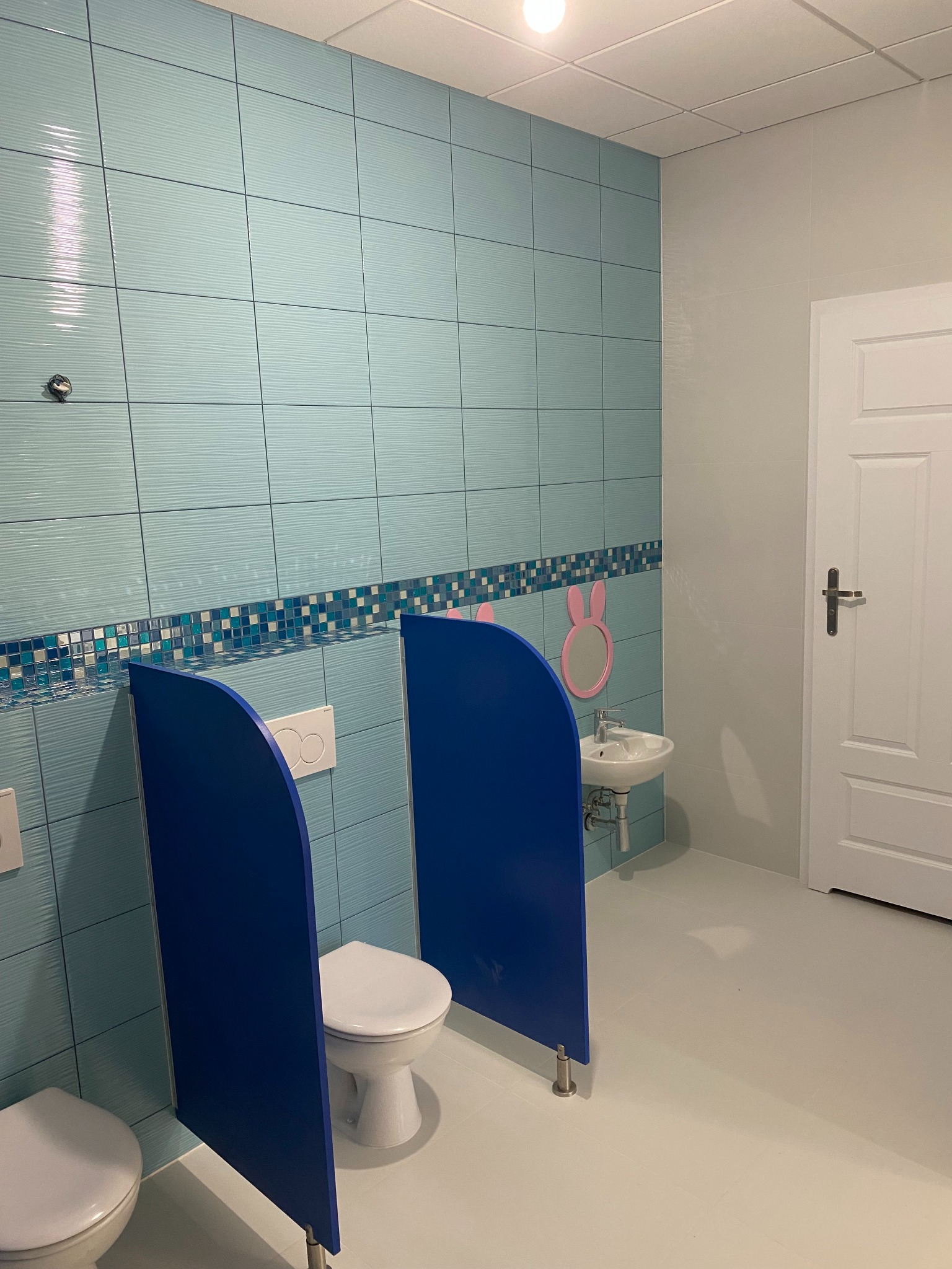 Jasne, błękitne płytki ścienne z dekoracyjnym pasem mozaiki w toalecie dla dzieci. Kabiny toaletowe oddzielone niebieskimi ściankami działowymi. Umywalka z lustrem w kształcie króliczka.