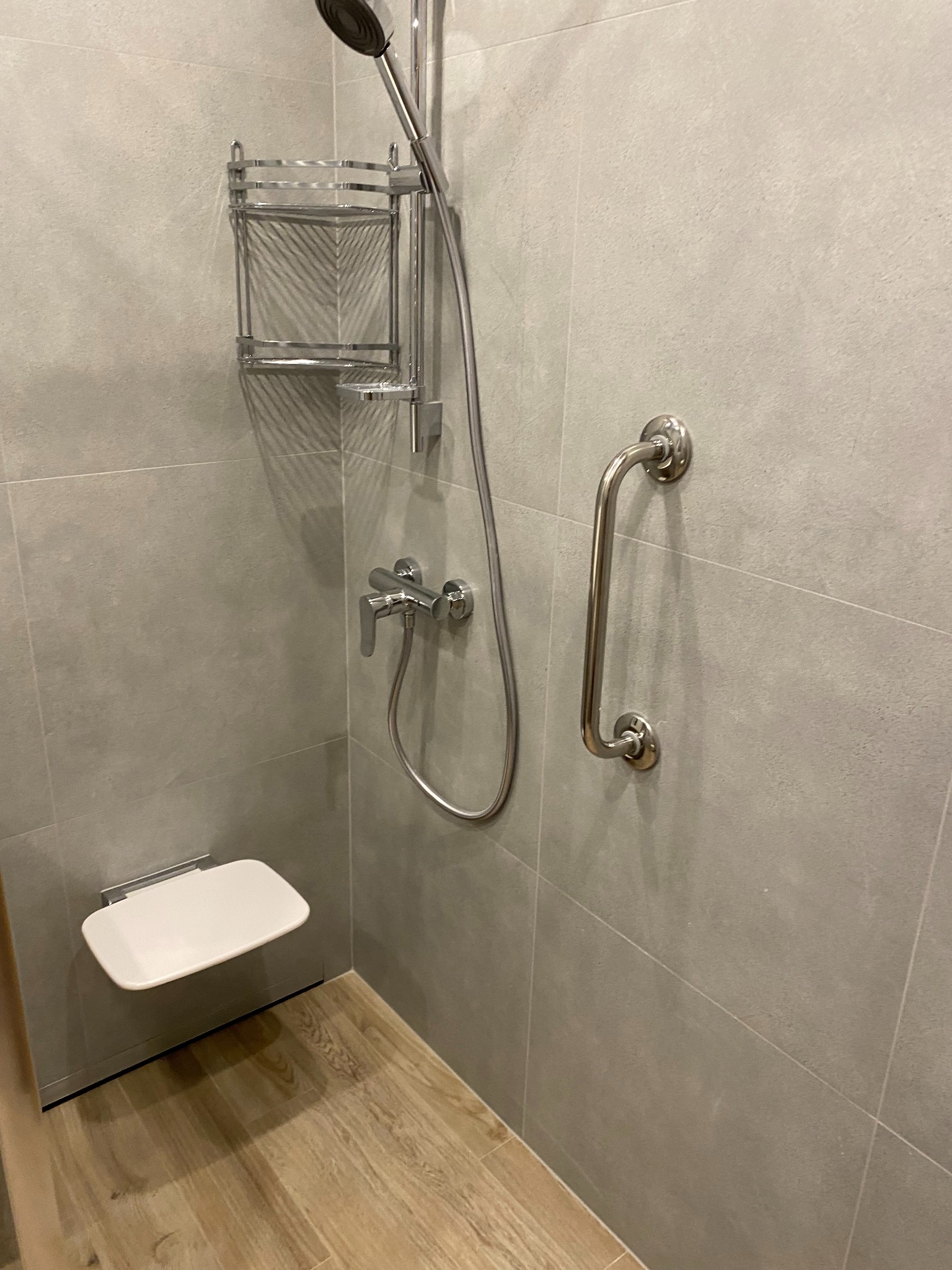Wykończona łazienka z szarymi płytkami, chromowaną armaturą, składanym siedziskiem i uchwytem dla niepełnosprawnych. Drewnopodobna podłoga. Minimalistyczny design.