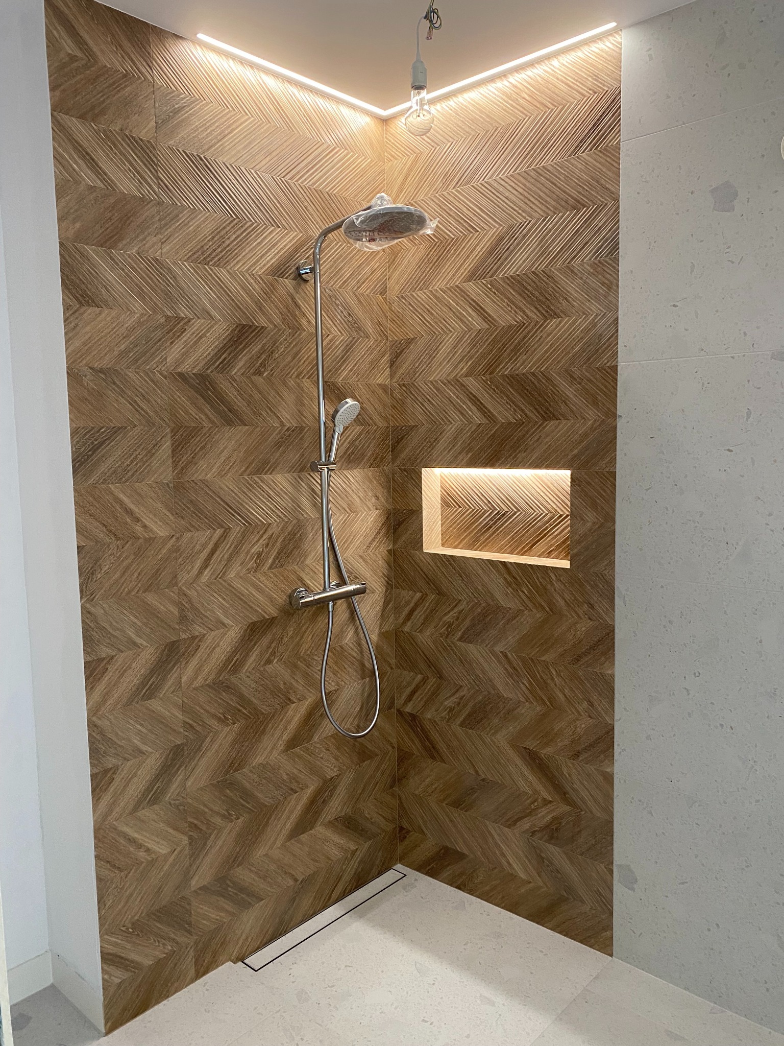 Nowoczesna kabina prysznicowa z drewnopodobną, wzorzystą okładziną ścienną i wnęką z oświetleniem LED. Chromowana armatura, minimalistyczny odpływ liniowy.