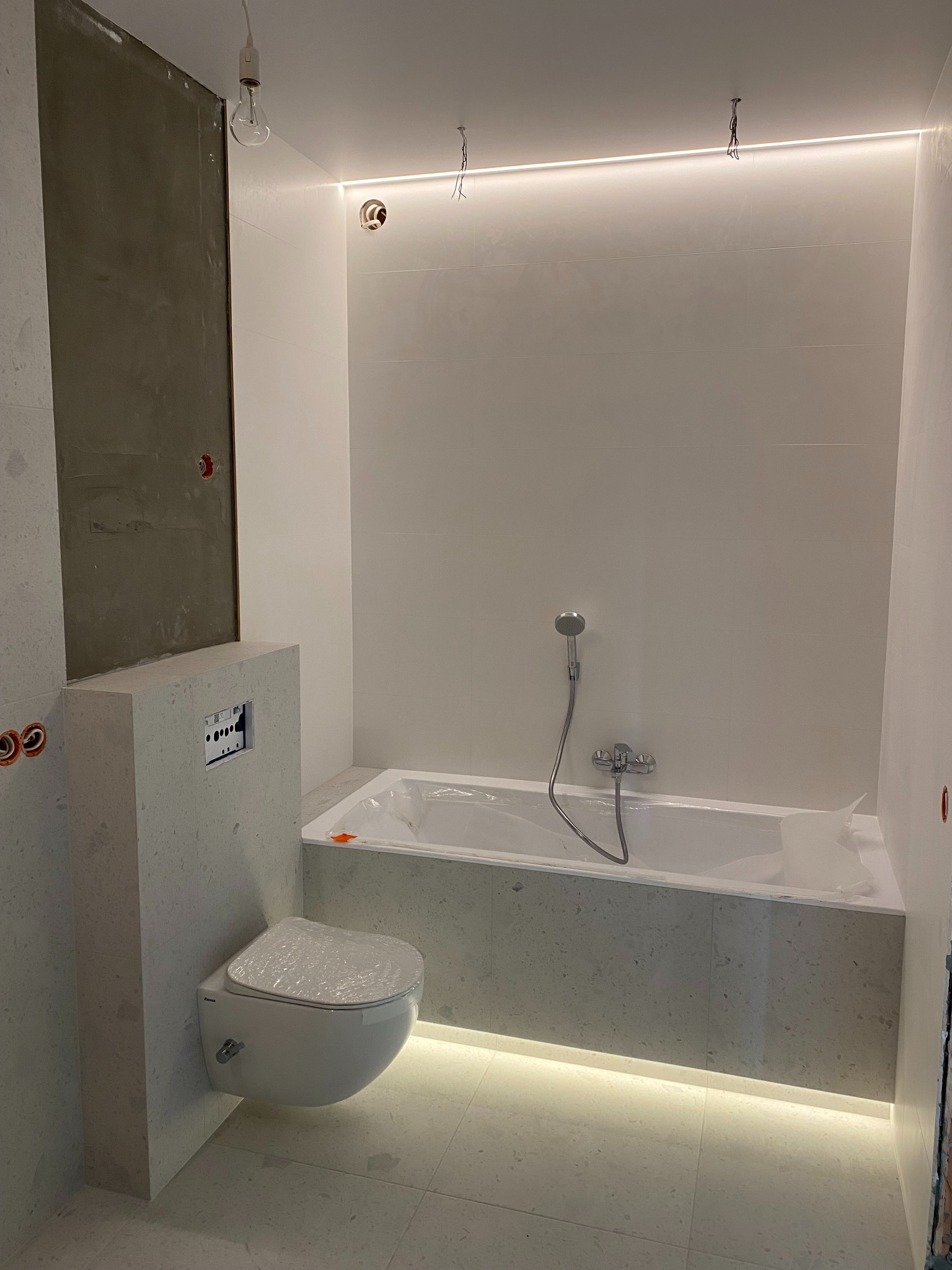 Remont łazienki: białe płytki, podwieszana toaleta z osłoną, wanna w folii, oświetlenie LED pod wanną i sufitowe. Częściowo wykończona ściana.