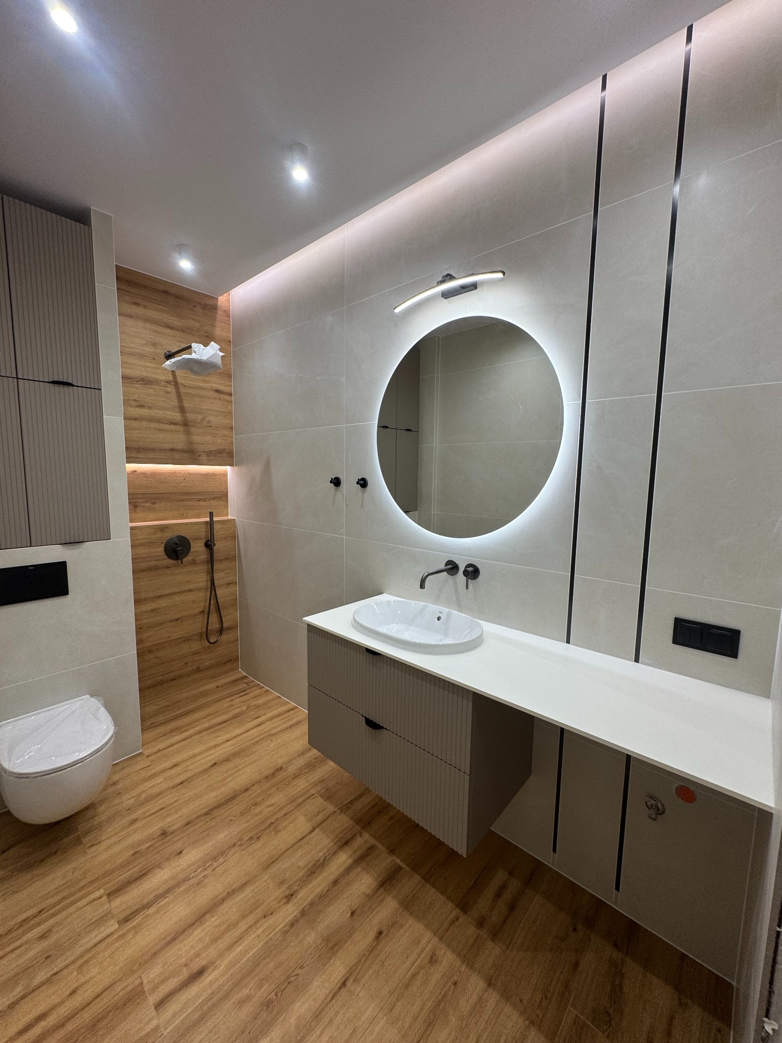 Nowoczesna łazienka po remoncie: okrągłe lustro z podświetleniem, drewnopodobne płytki, minimalistyczna armatura i szafki w odcieniach szarości.