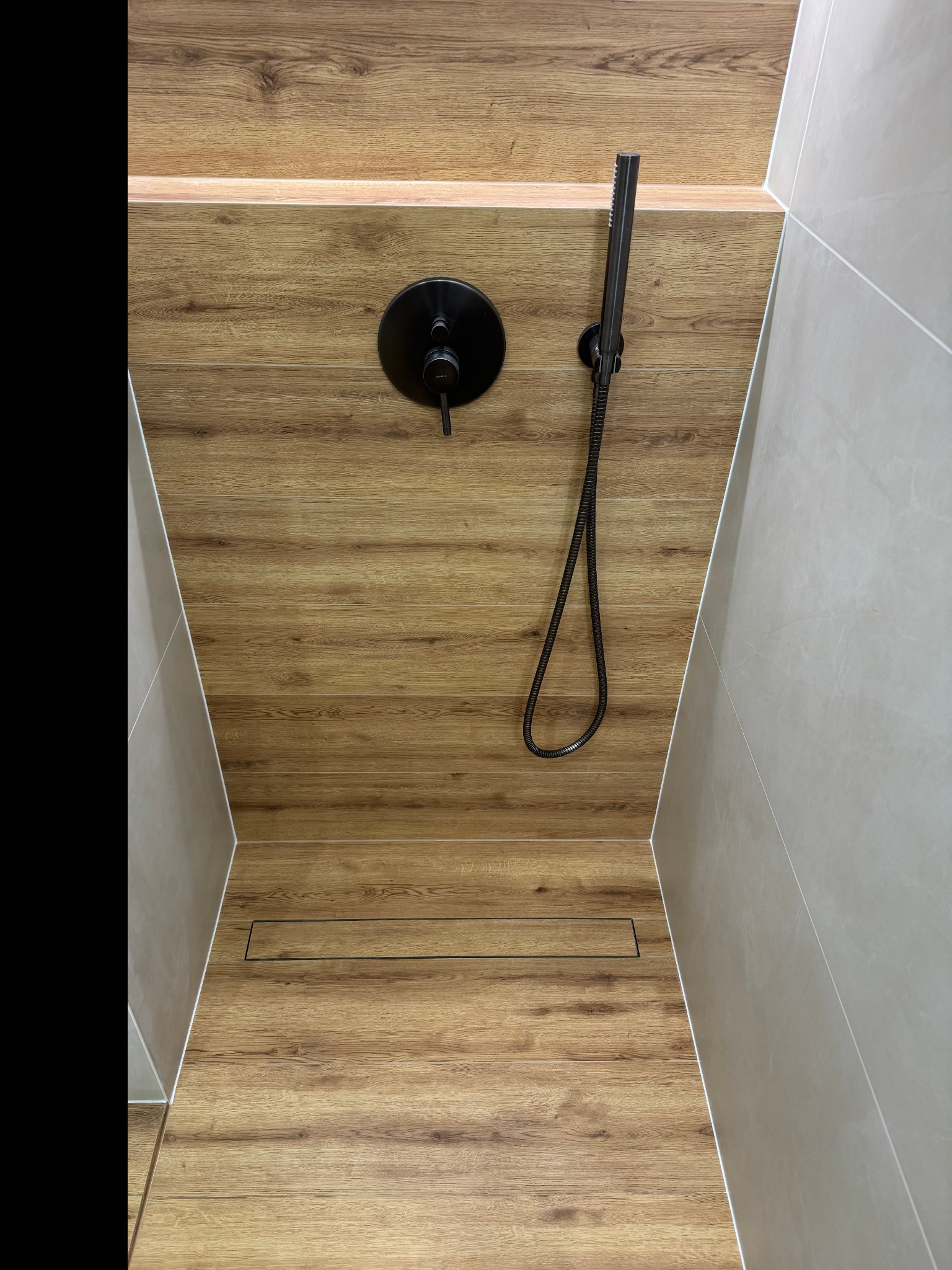 Nowoczesny prysznic walk-in z drewnopodobną okładziną, czarną armaturą i odpływem liniowym. Minimalistyczny design, jasne płytki na ścianach.